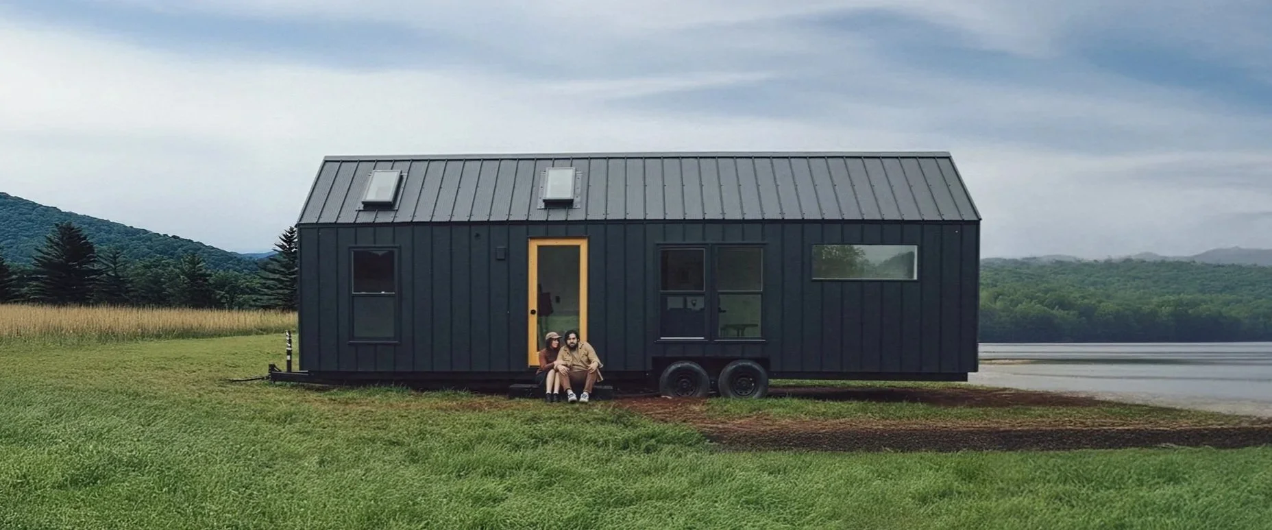 A modern black tiny house on wheels with a yellow door and several windows, situated on a grassy field with trees and hills in the background. Two people sit on the steps near the door.