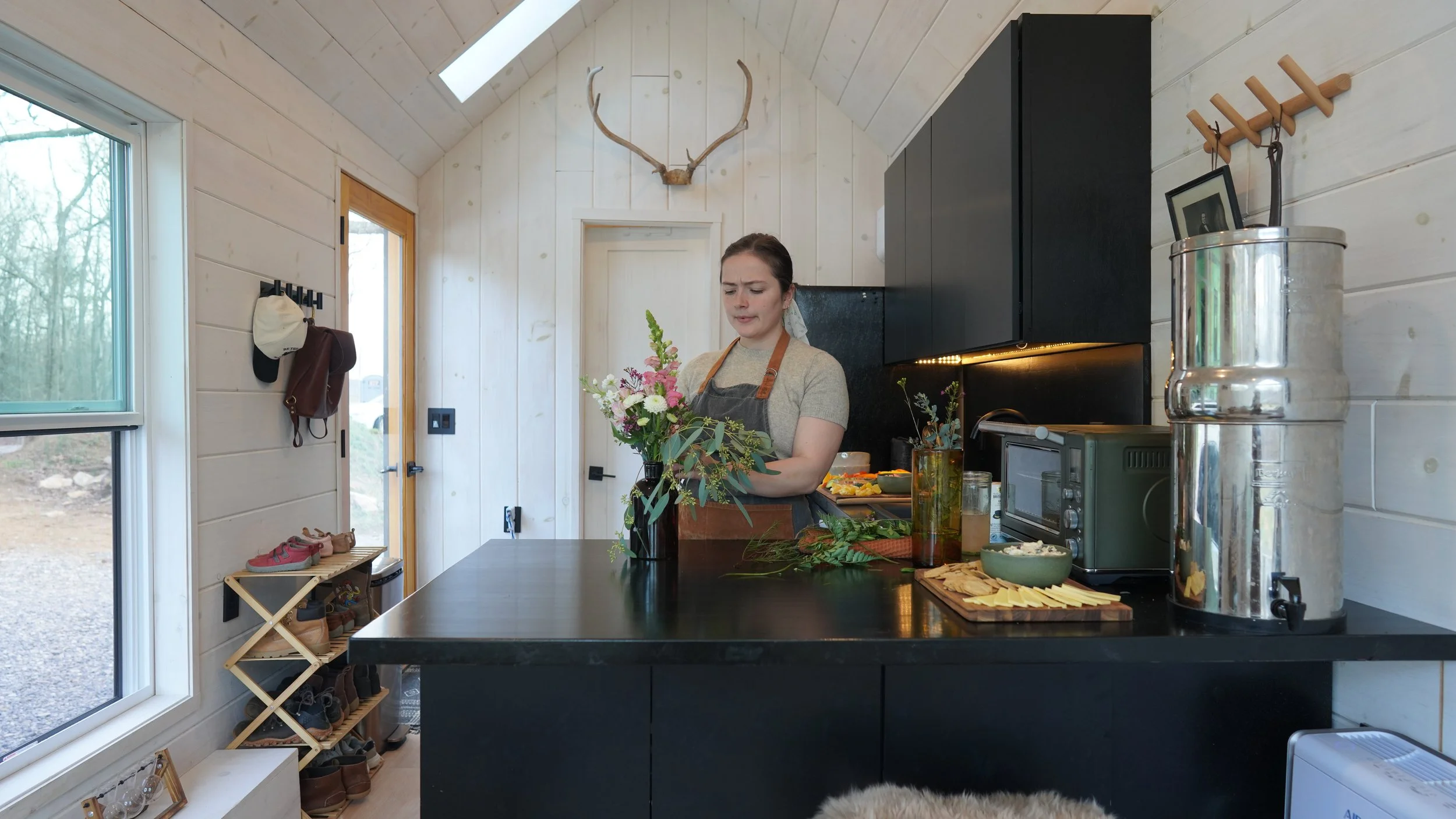 A woman in a gray shirt and apron stands in a modern rustic kitchen, arranging a bouquet of flowers on a black countertop. The kitchen has light wood-paneled walls, a deer antler decoration above the door, and a large window to the outside. Various kitchen items, including a toaster oven, a water dispenser, and a cutting board with cheese, are visible.