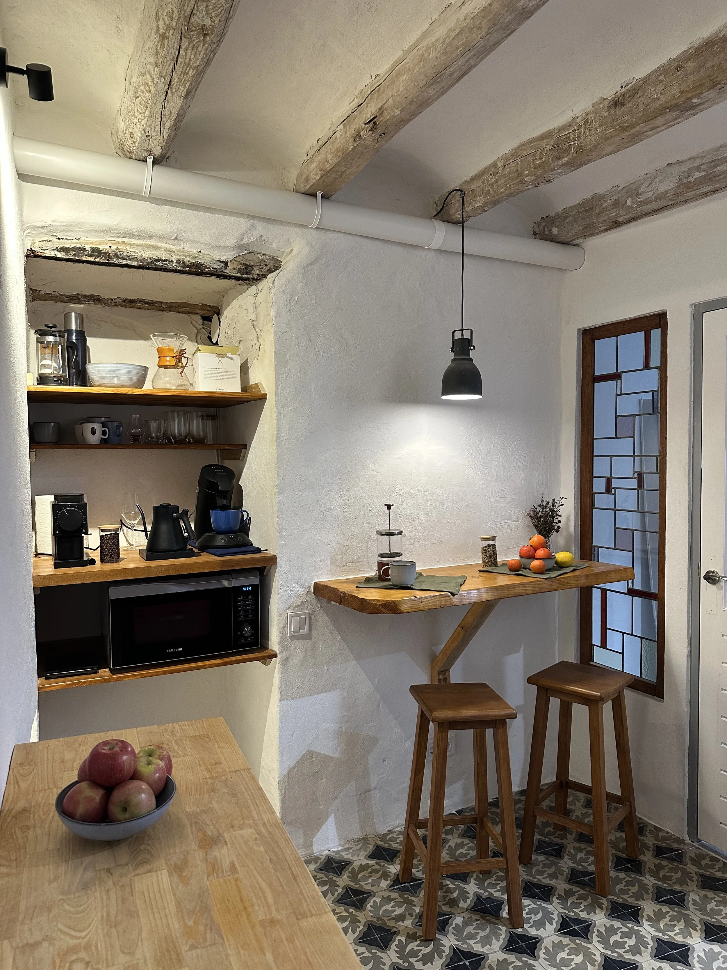 Cozy kitchen corner with wooden shelves, a microwave, coffee maker, French press, and various kitchen items. A small wooden bar with two stools and a hanging light. Apples in a bowl on the counter. Rustic beams and decorative tile floor.