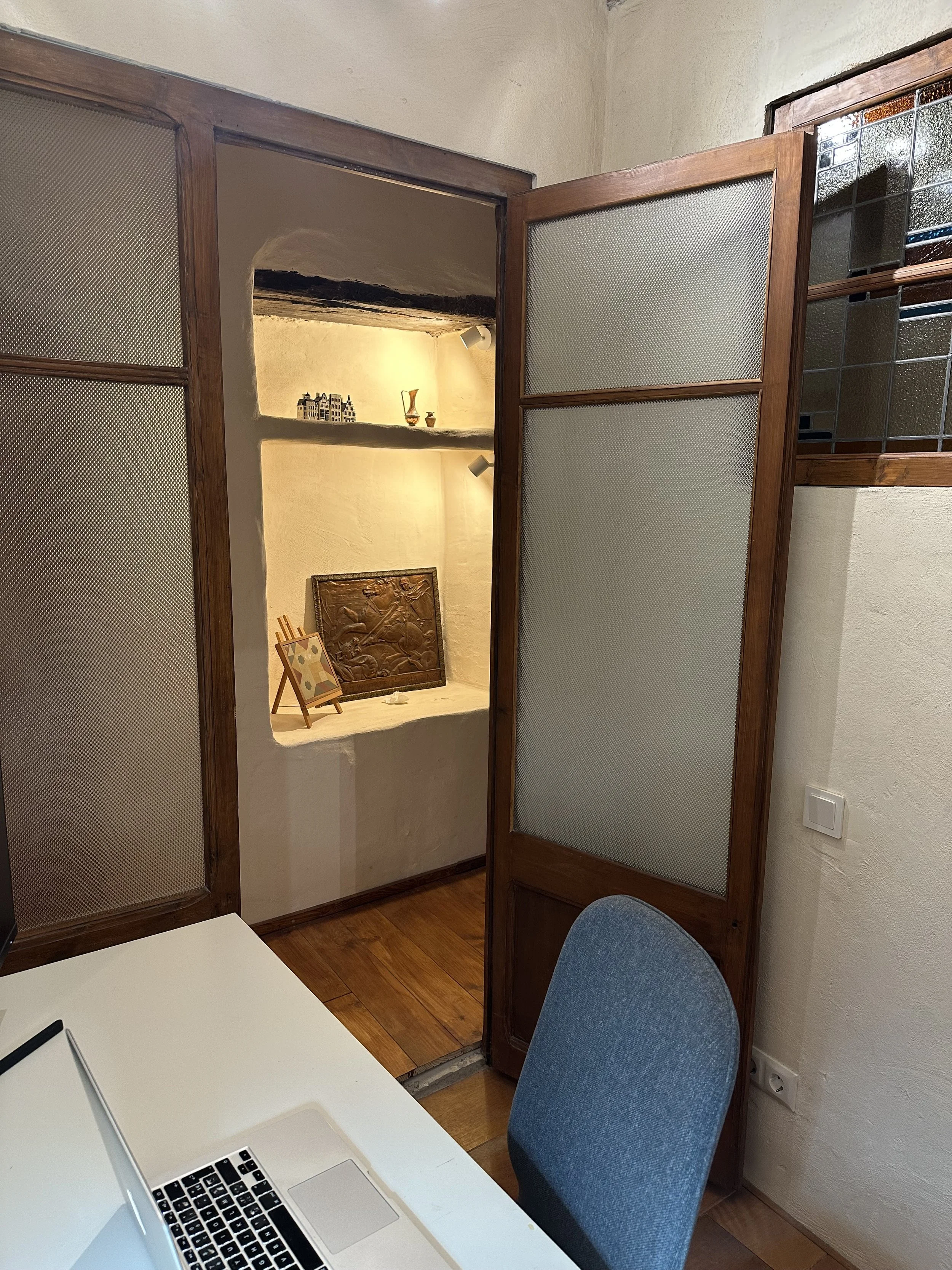 Office with desk, laptop, chair, open door with shelves displaying decorative items, wooden floor, and textured walls.