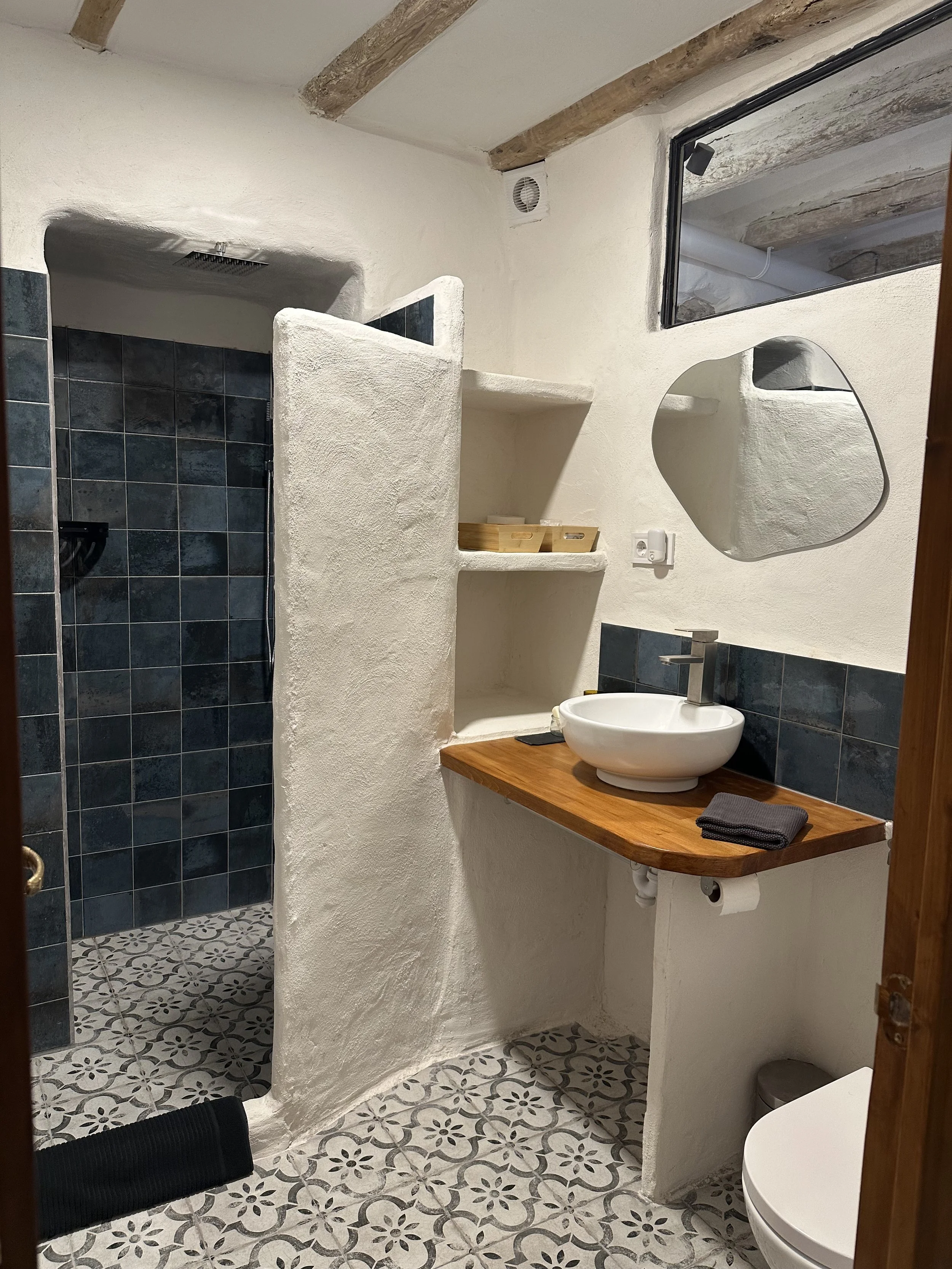Modern bathroom with rustic design featuring white stucco walls, wooden beams, tiled shower with gray tiles, curved wooden countertop with a white vessel sink, circular mirror, and patterned floor tiles.