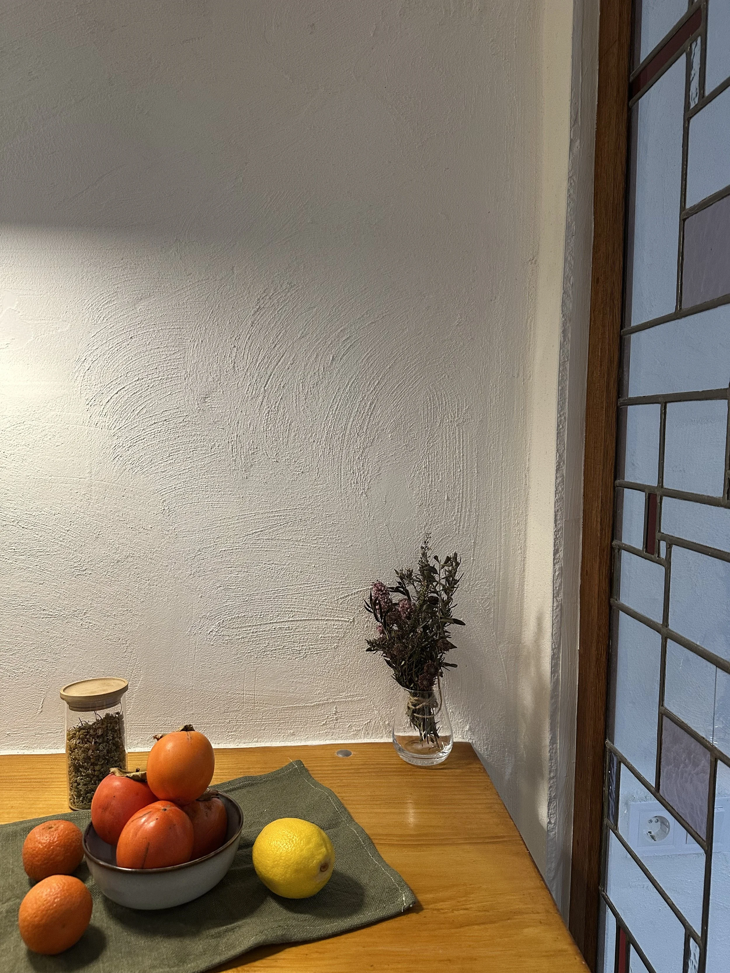 Wooden table with a bowl of fruit including persimmons, a lemon, and tangerines, a jar with dried flowers, and a small vase with a dried bouquet near a stained glass window.