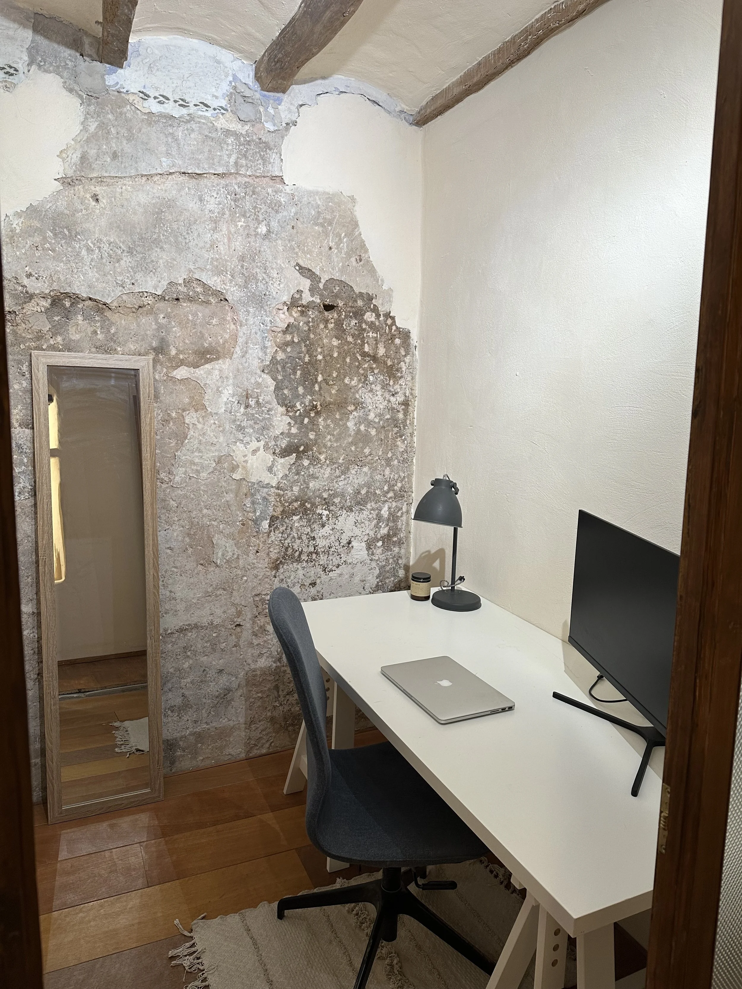 Small home office with a rustic vibe, featuring a white desk, laptop, monitor, desk lamp, gray office chair, wooden floor, and a distressed wall with exposed plaster. A full-length mirror leans against the wall, enhancing the minimalist decor.