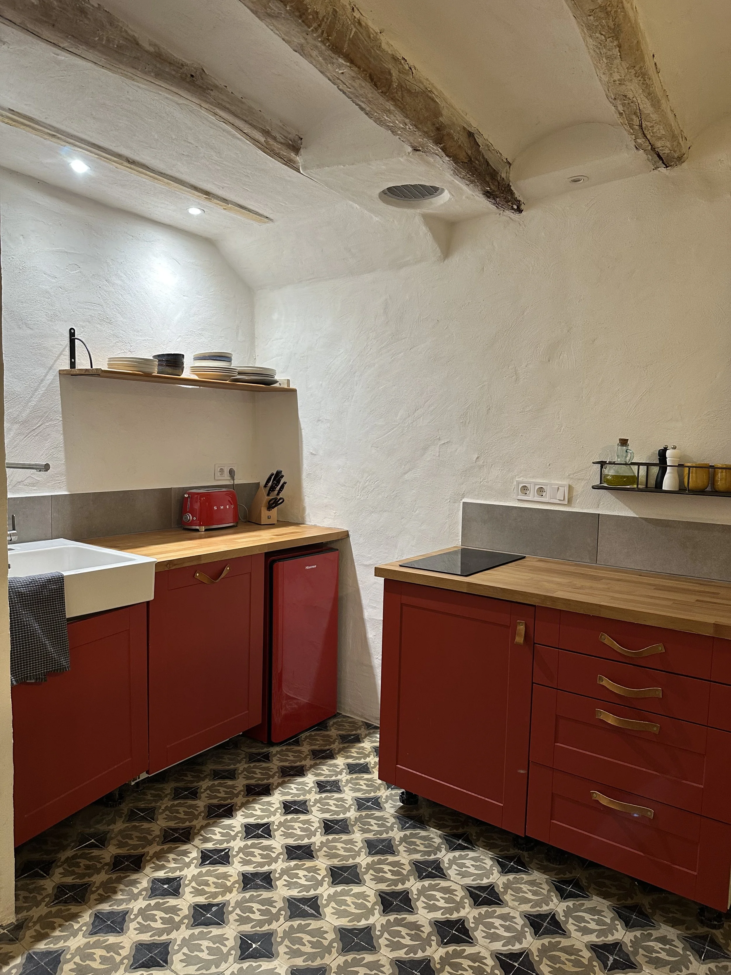 Cozy kitchen with red cabinets, wooden countertops, a white farmhouse sink, stove, red mini fridge, and decorative floor tiles.