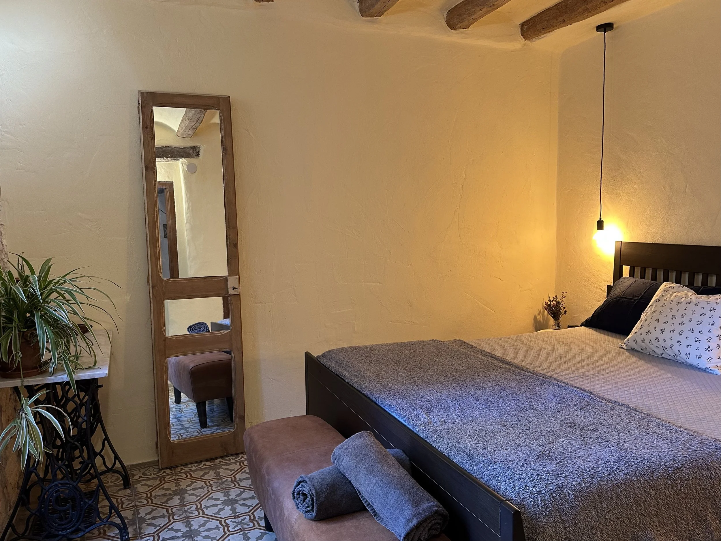 Cozy bedroom with a wooden bed, soft bedding, a hanging light, a standing mirror, potted plant, and a small bench with rolled towels.
