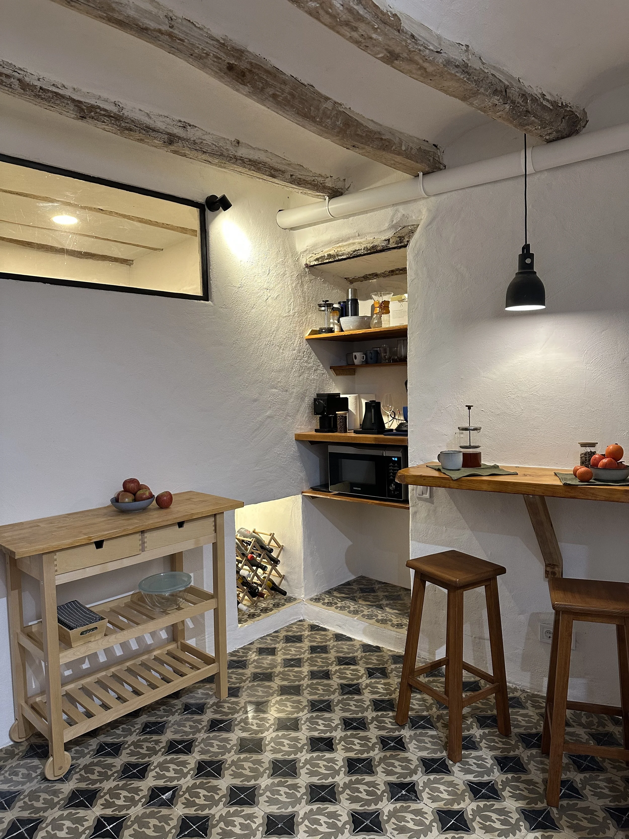 Cozy kitchen with tile floor, wooden beams, and a small breakfast bar with stools. Includes a rolling table with apples, corner shelves with kitchenware, and a wine rack.