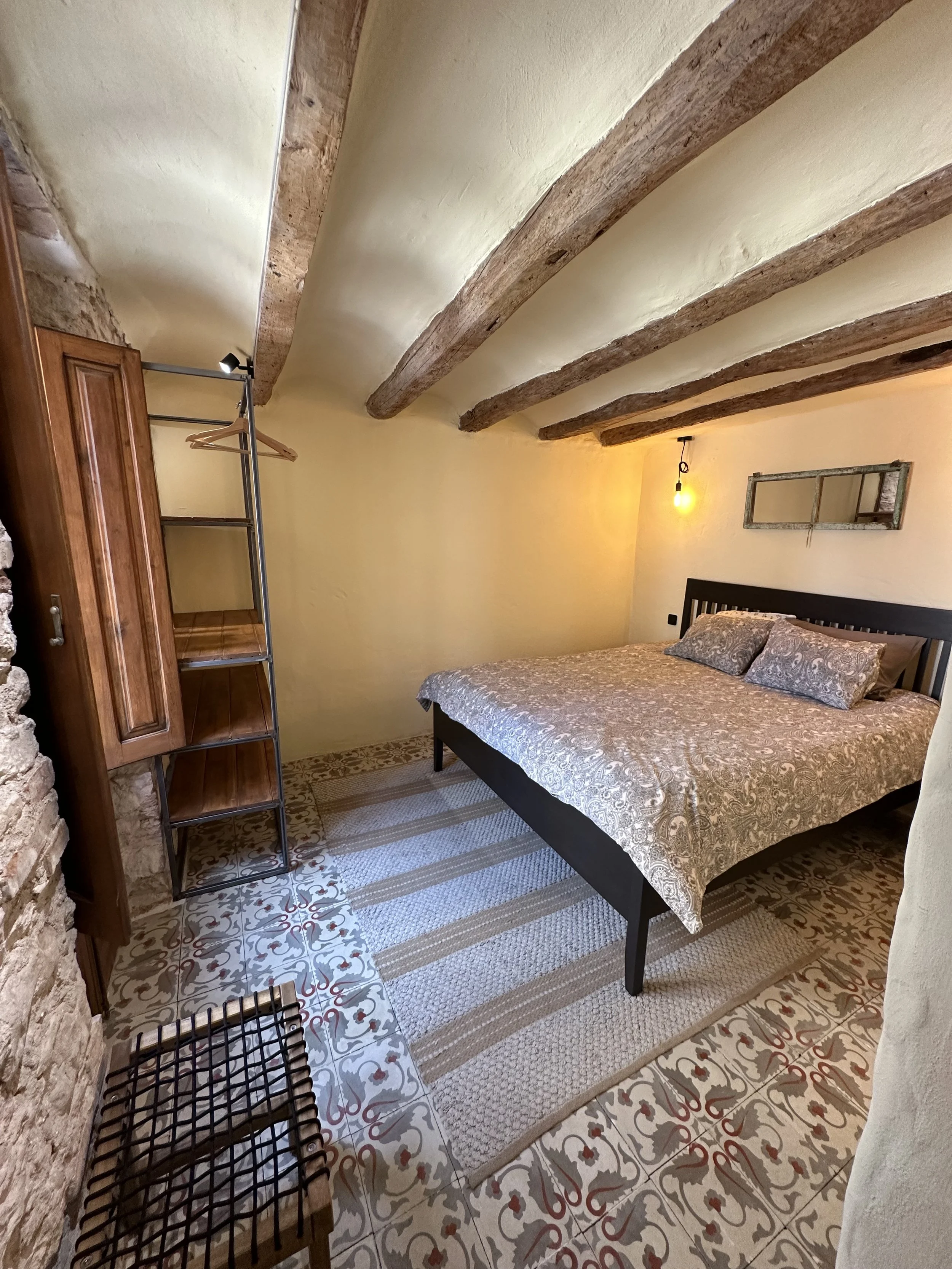 Rustic bedroom with wooden beams, a large bed with patterned bedding, decorative tiled floor, and a hanging light. There is a wooden wardrobe with shelves and colorful carpet runner.