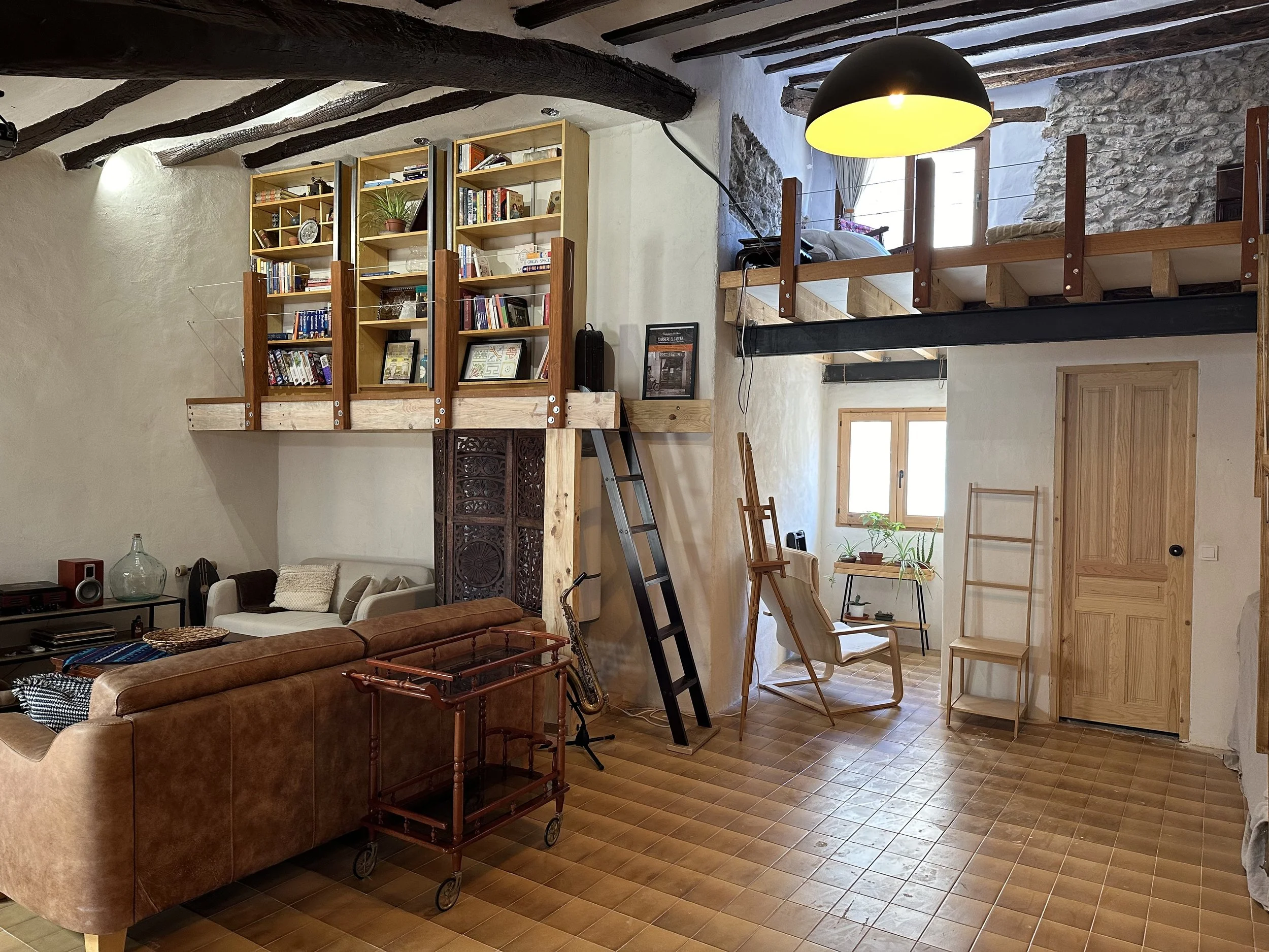 Loft-style living room with wooden beams, bookshelves, a brown sofa, ladder, easel, and a loft bed.