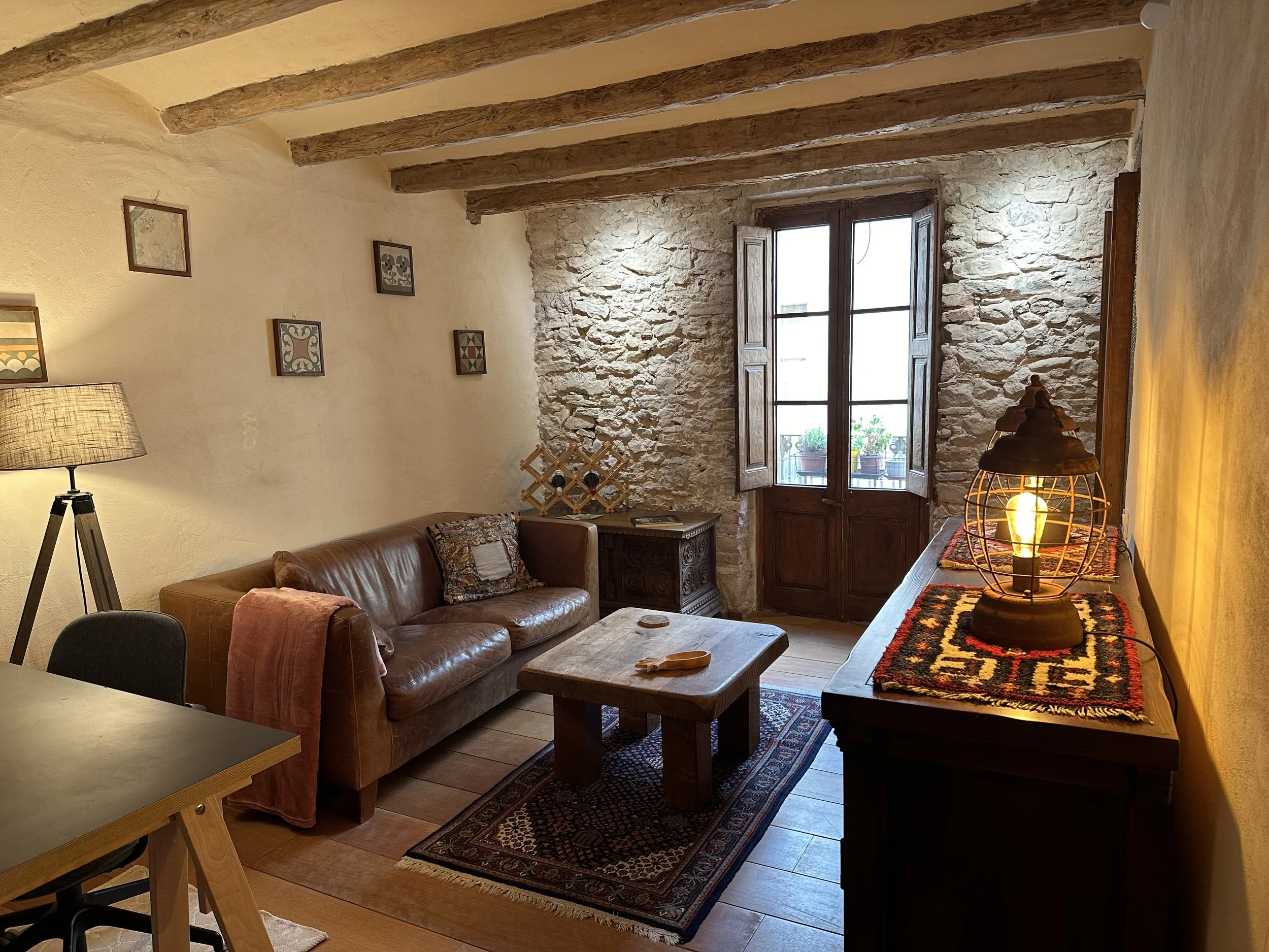 Cozy living room with a leather couch, wooden coffee table, and rustic decor. Exposed wooden beams on the ceiling, a stone wall, and a window with wooden shutters. A decorative lamp sits on a wooden sideboard, and framed art hangs on the wall.