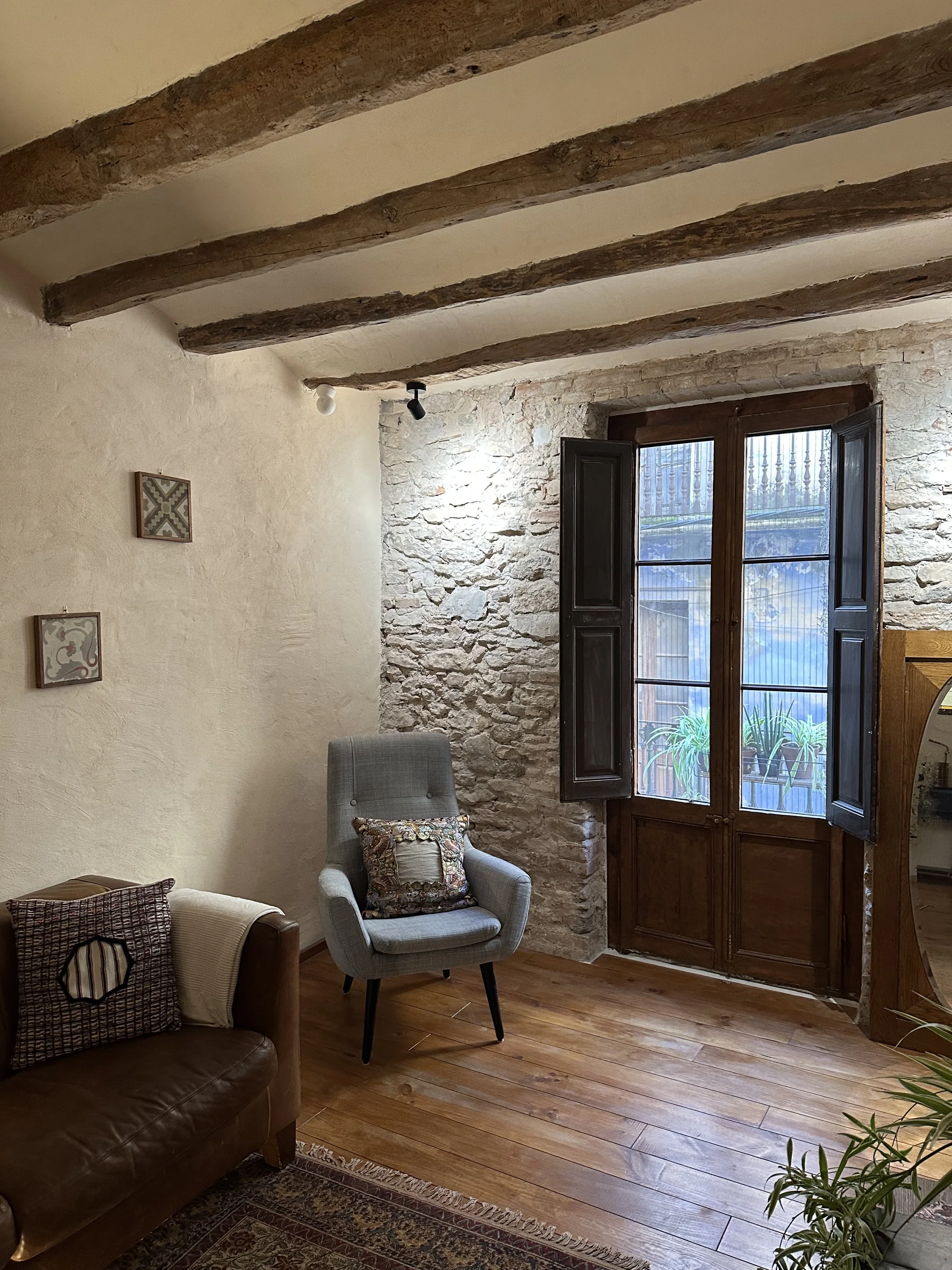 Cozy living room with exposed wooden beams, a stone wall, and a large wooden-framed window. The room features a leather sofa with decorative pillows, a gray upholstered armchair, a decorative rug, and plants outside the window.