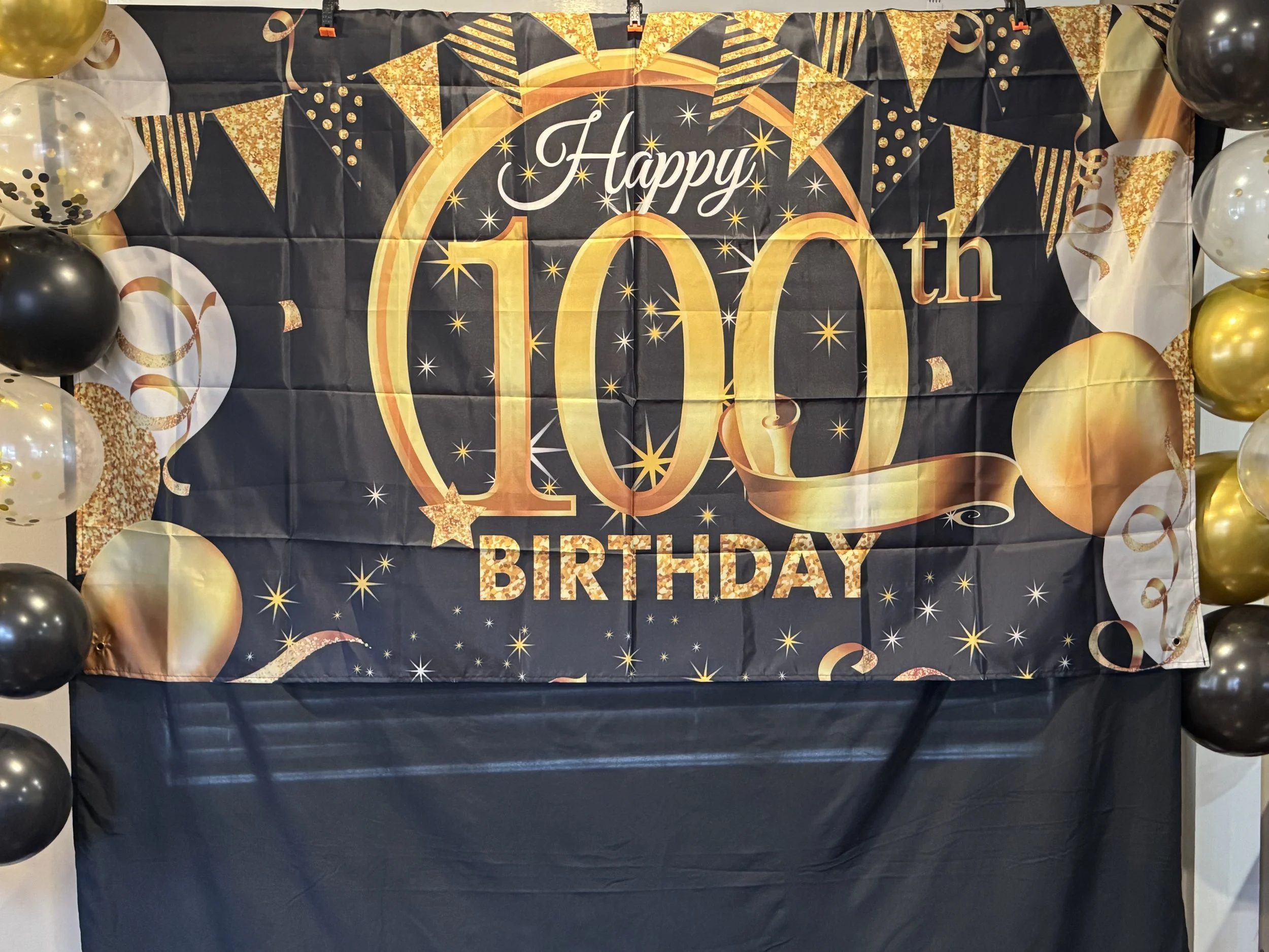 Decorative backdrop for a 10th birthday celebration with black, gold, and white balloons framing a black banner featuring gold and white text and designs that reads "Happy 10th Birthday" with stars and bunting graphics.