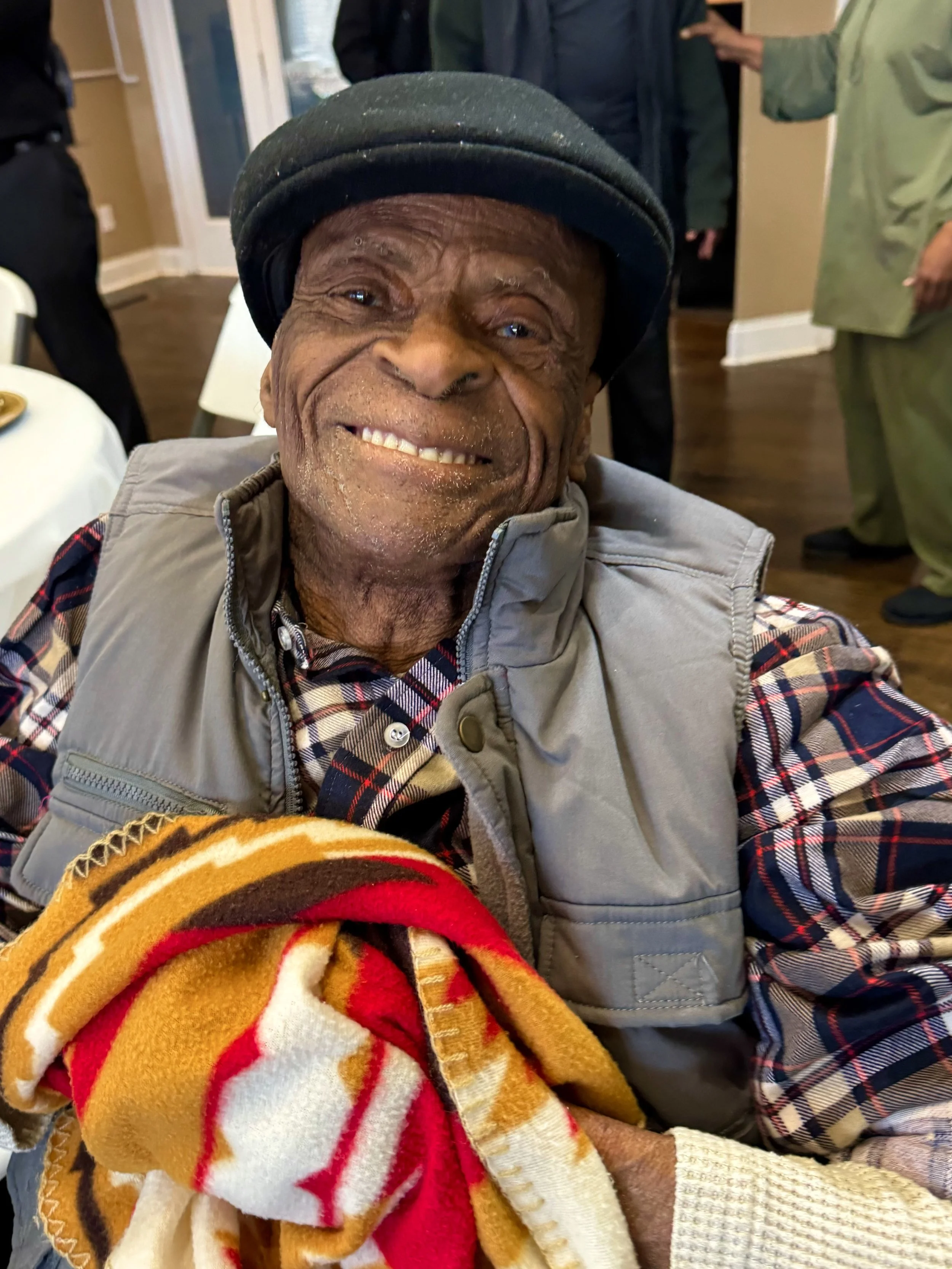 Smiling elderly man wearing a black hat, plaid shirt, and a gray vest, holding a colorful blanket, in a room with other people in the background.