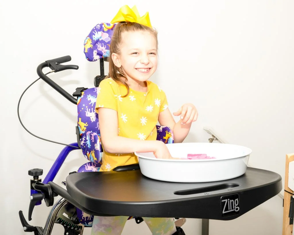 Young girl in a bright yellow t-shirt in a standing frame playing with a sensory tray.