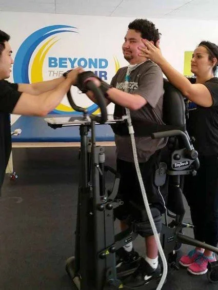 Teenage boy being helped by two physiotherapists with a standing frame in a gym with bright blue and yellow signage.