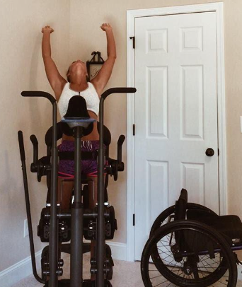 Female in gym wear on a standing frame in her front room - she's raising arms above her head to stretch.