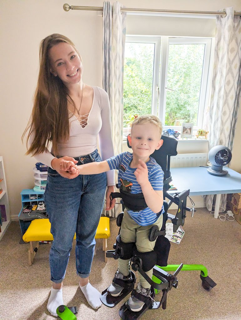 Young boy in an easystand frame with his mum holding his hand beside him