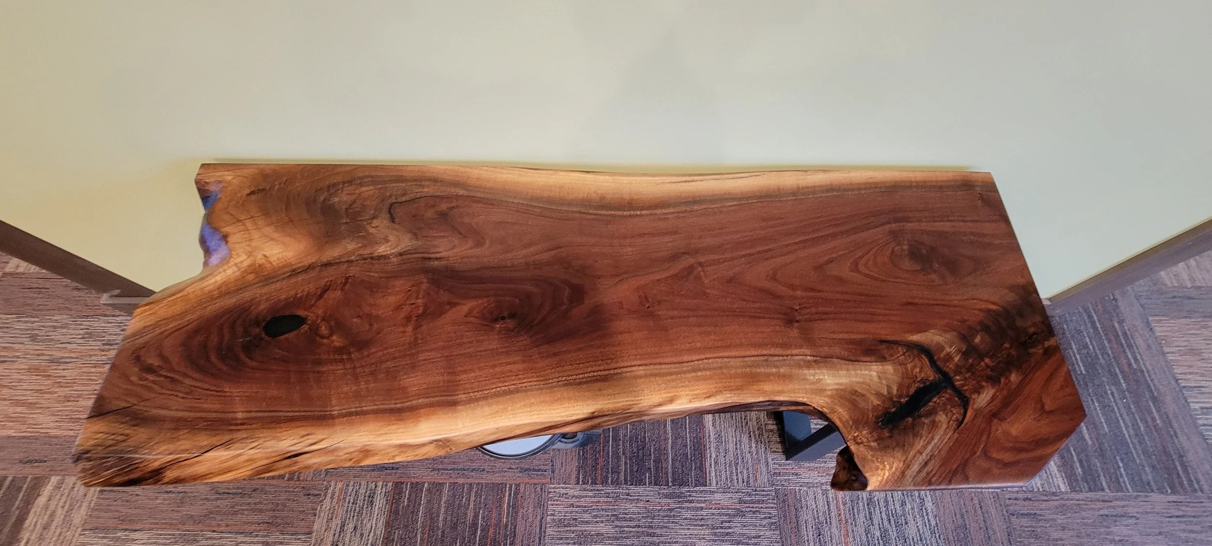 Close-up of a polished, natural wood table with a live edge, showing the wood grain and knots, placed on a patterned carpet with a light green wall in the background.