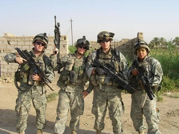 Four soldiers in camouflage uniforms standing outdoors with weapons, in a desert-like environment with some low walls and sparse greenery.