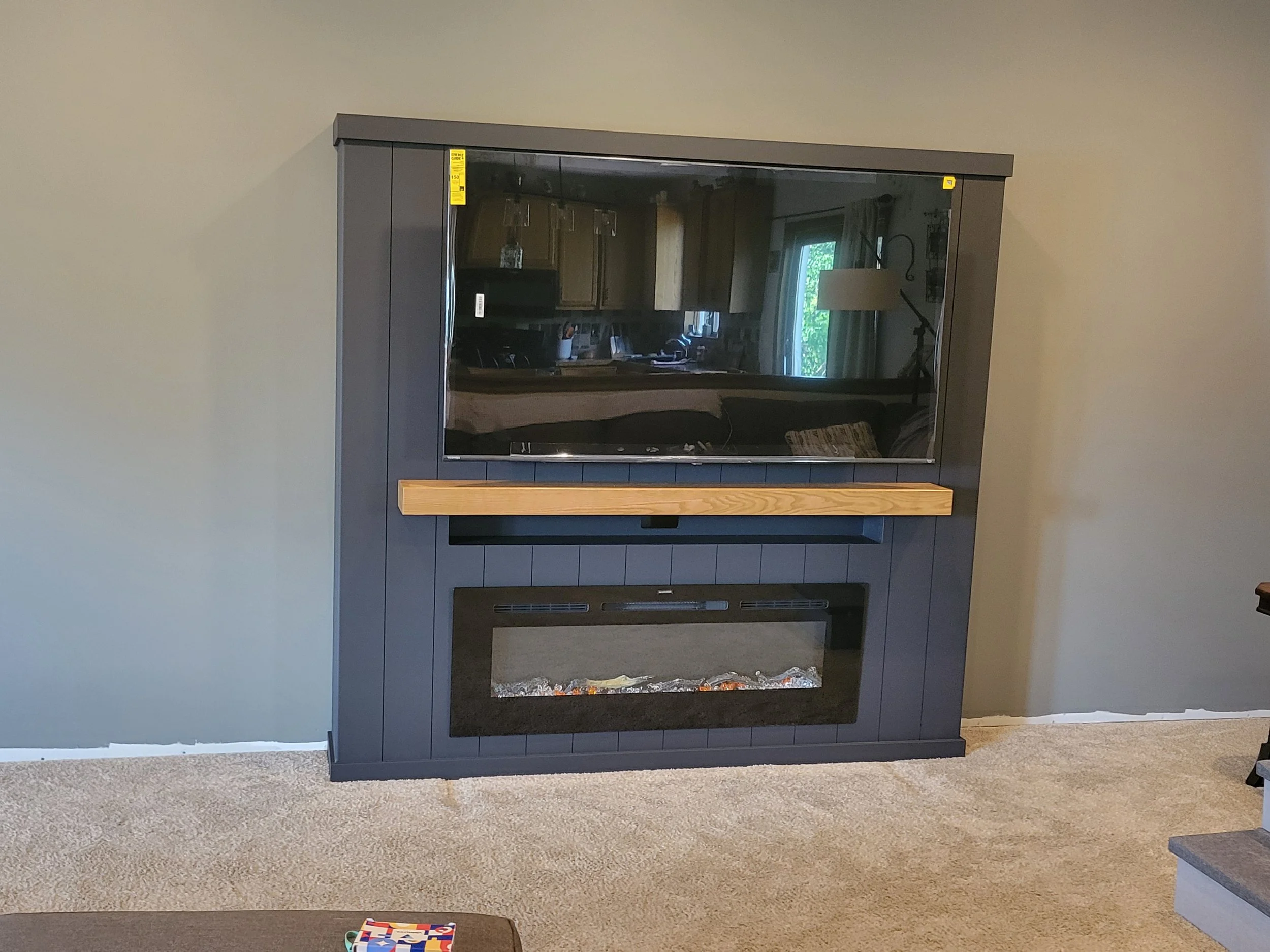 A modern dark gray fireplace with a glass front, built into a light beige wall. The fireplace has a glowing fire and a strip of wood above the mantel. The room has a beige carpet and a window with curtains reflected in the glass.