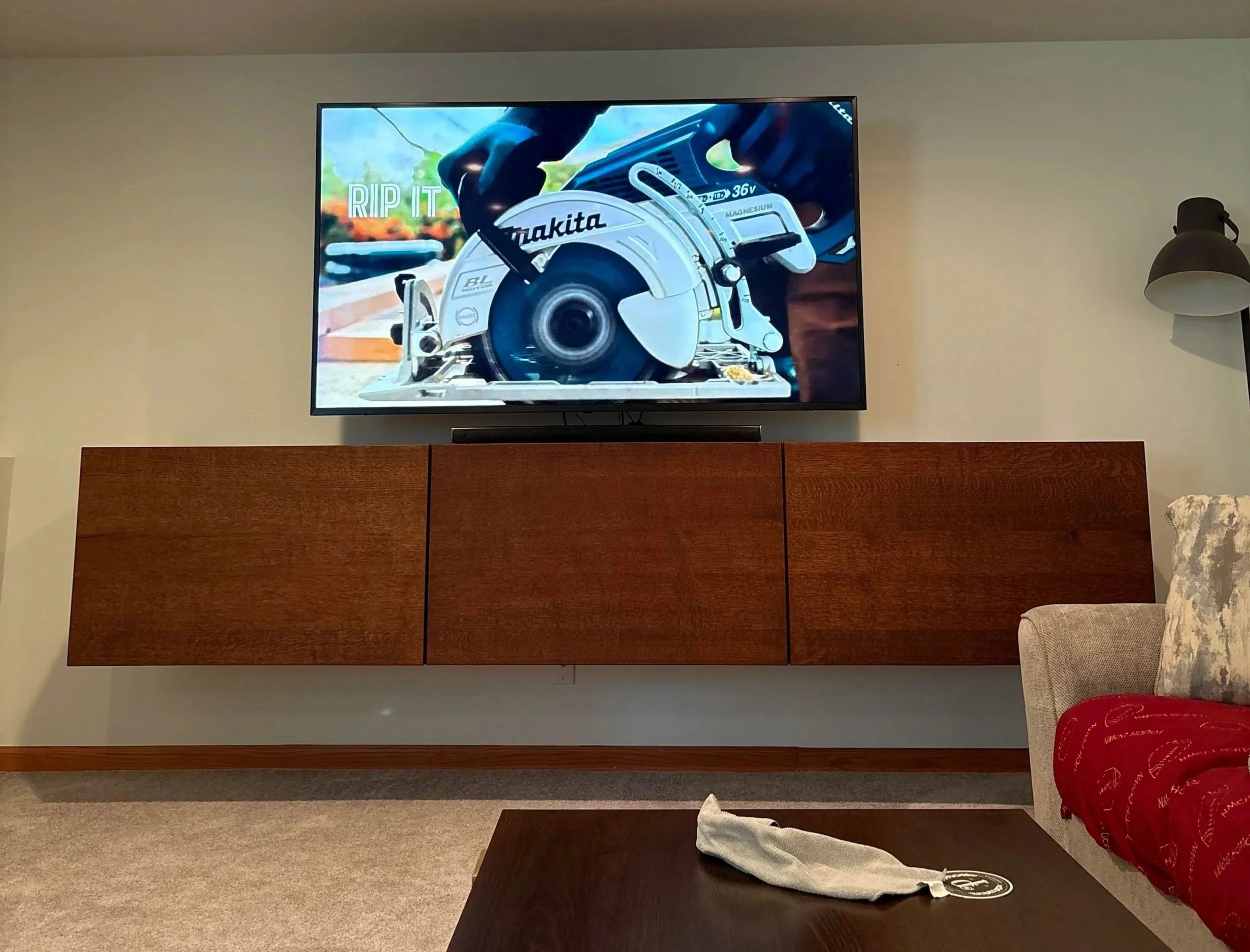 Living room with a flat-screen TV mounted on the wall displaying a construction saw with the text "RIP IT". There is a brown cabinet below the TV. To the right, part of a beige and yellow patterned armchair and a red blanket on the armchair are visible. To the right of the TV, a wall-mounted lamp is on. The room has beige walls and a carpeted floor.