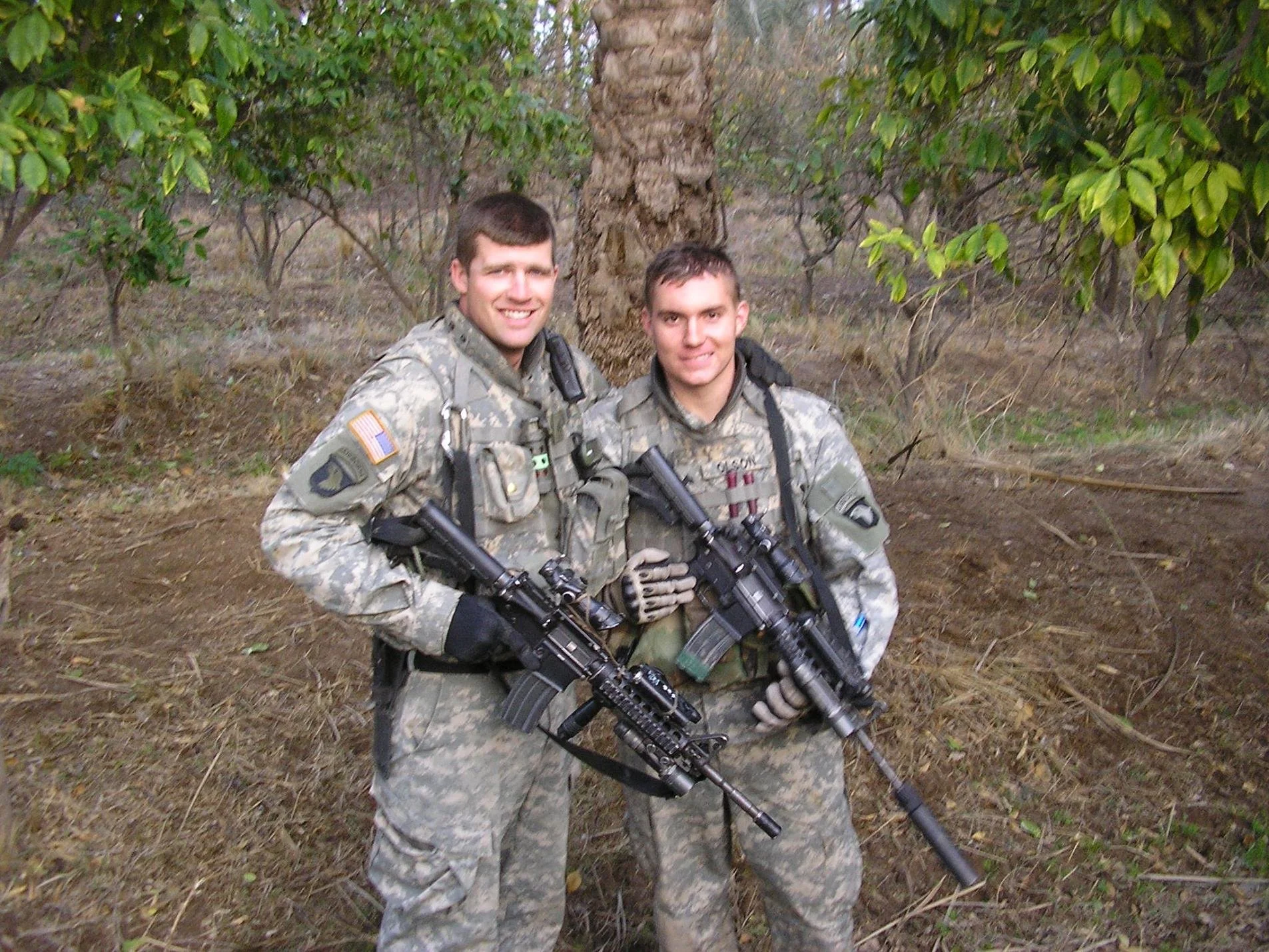 Two soldiers in camouflage uniforms standing in front of a tree in a wooded area, holding rifles.