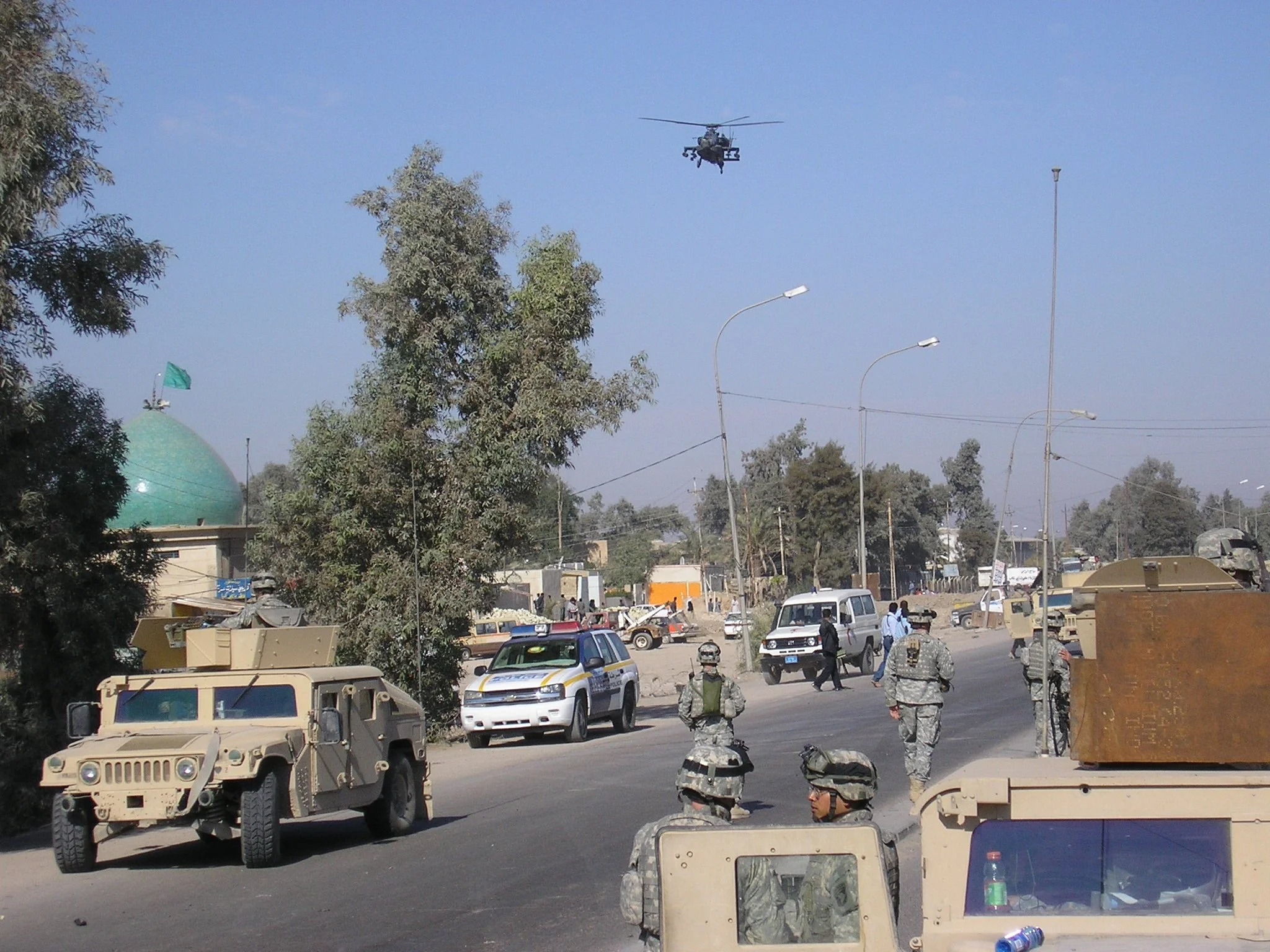 Military vehicles and soldiers on a busy street with trees, streetlights, and a helicopter in the sky.