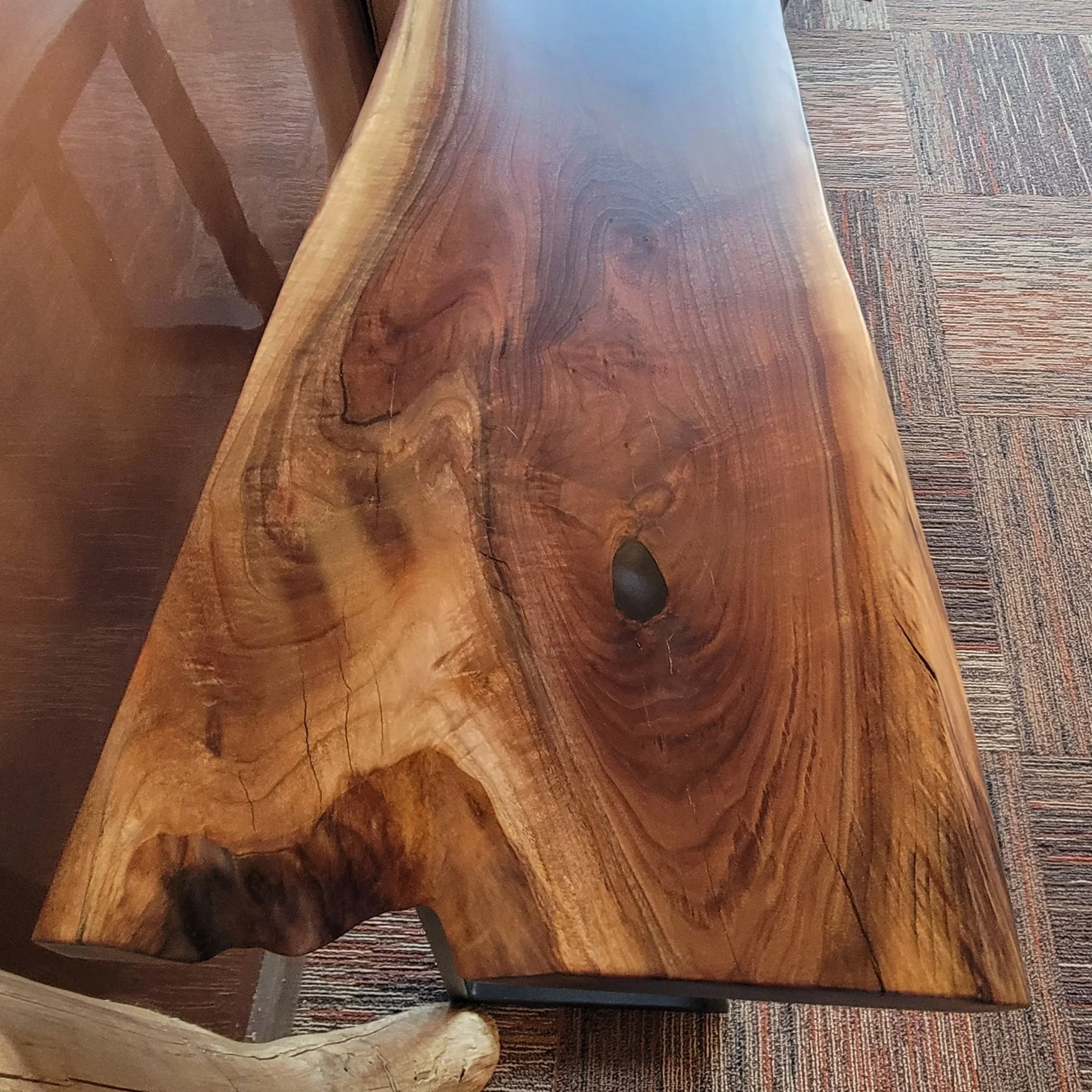 Close-up of a wooden bench with a natural, polished finish, showing the wood grain and knots, placed on a patterned carpeted floor.