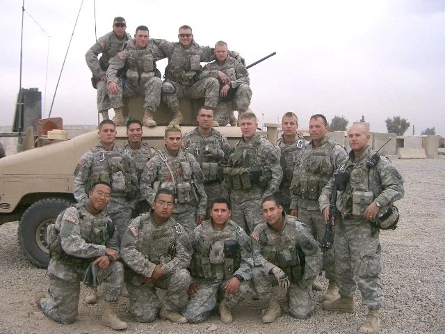 Group of soldiers in uniform posing outdoors with military vehicles in a desert setting.