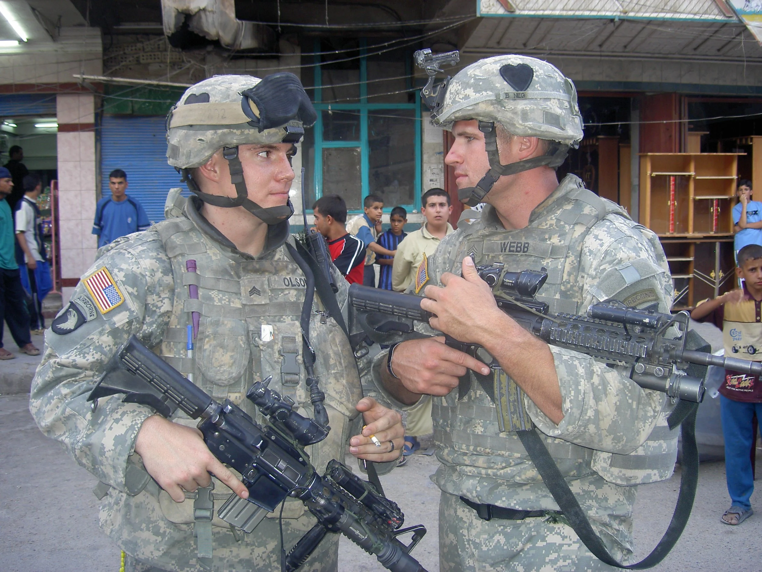 Two soldiers in camouflage uniforms and helmets shaking hands on a busy street, surrounded by onlookers and children.