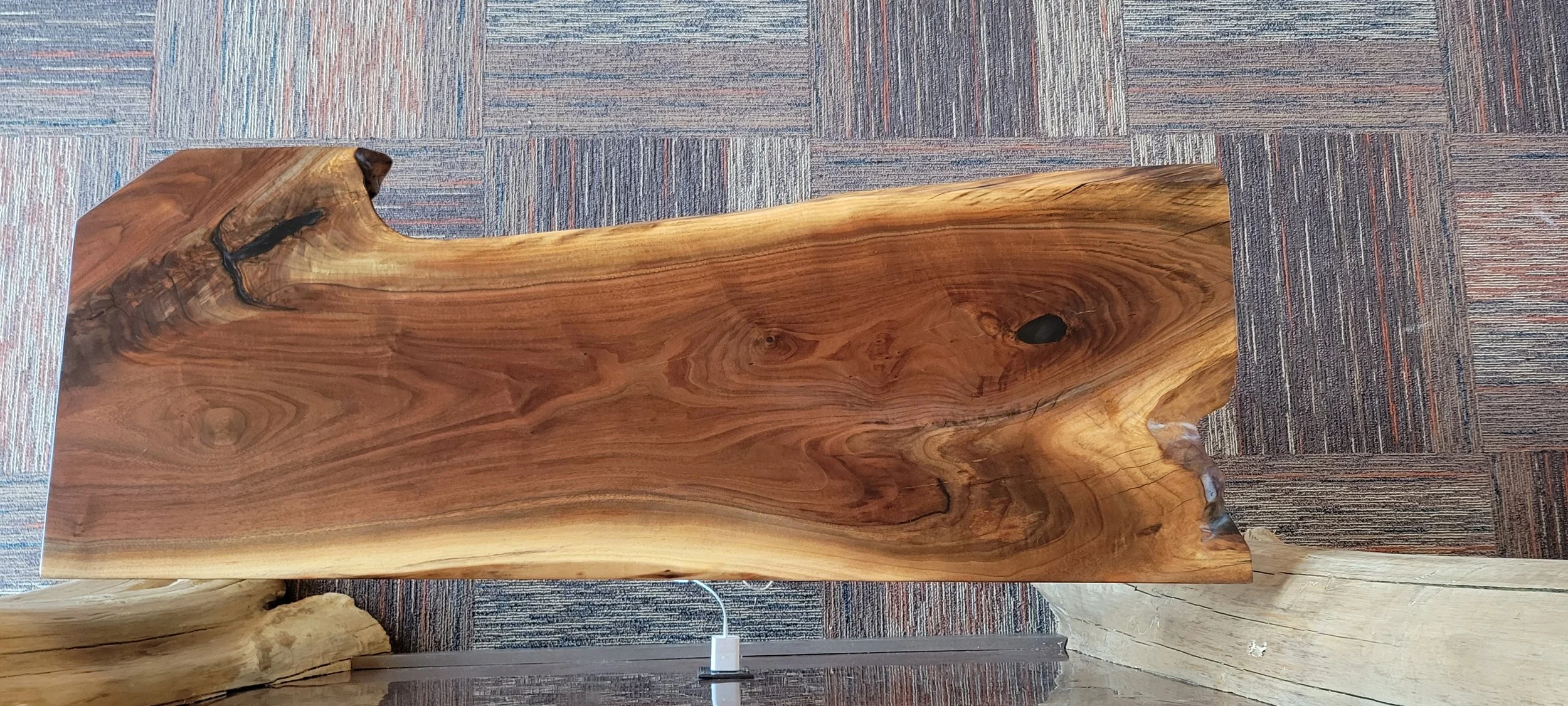 A large, polished, natural wood slab with visible grain patterns and knots, displayed indoors on a carpeted floor.