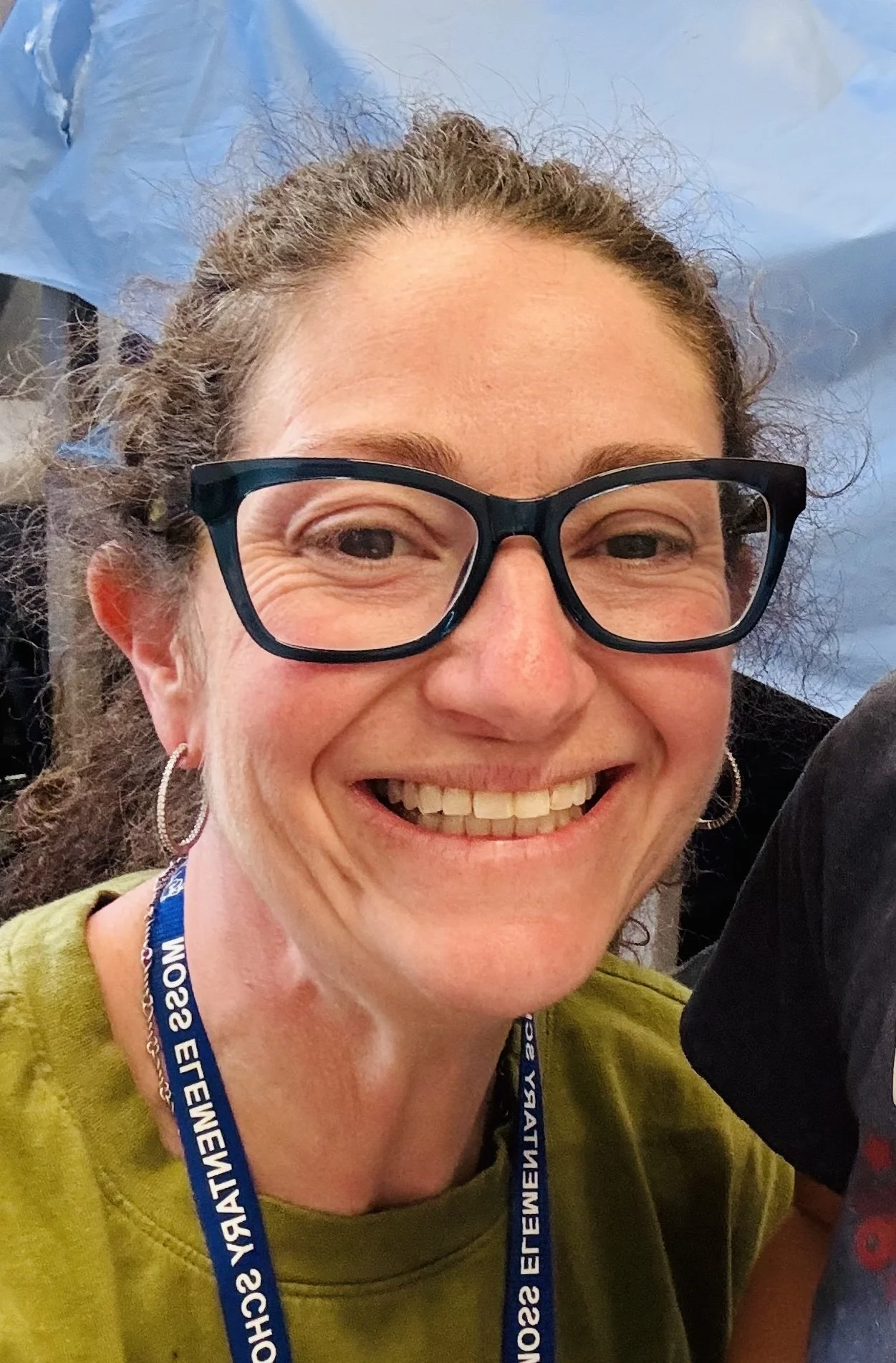 A woman with glasses smiling at the camera, wearing an event lanyard, with a blue sky in the background.