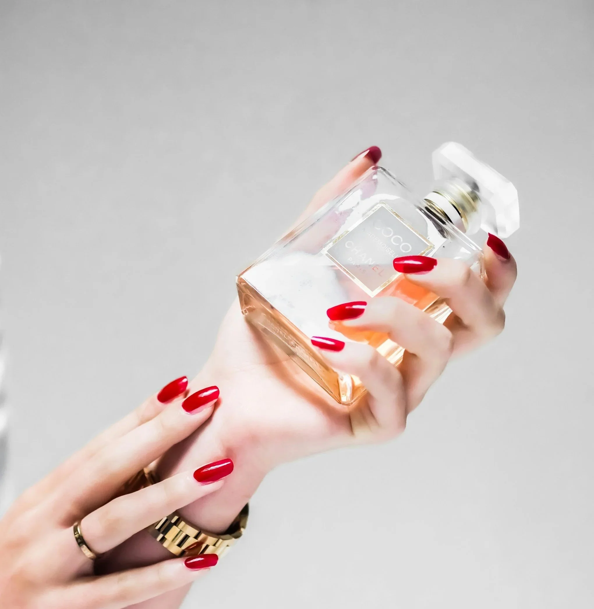 Hand holding a bottle of Chanel Coco Mademoiselle perfume with manicured red nails and a gold watch.