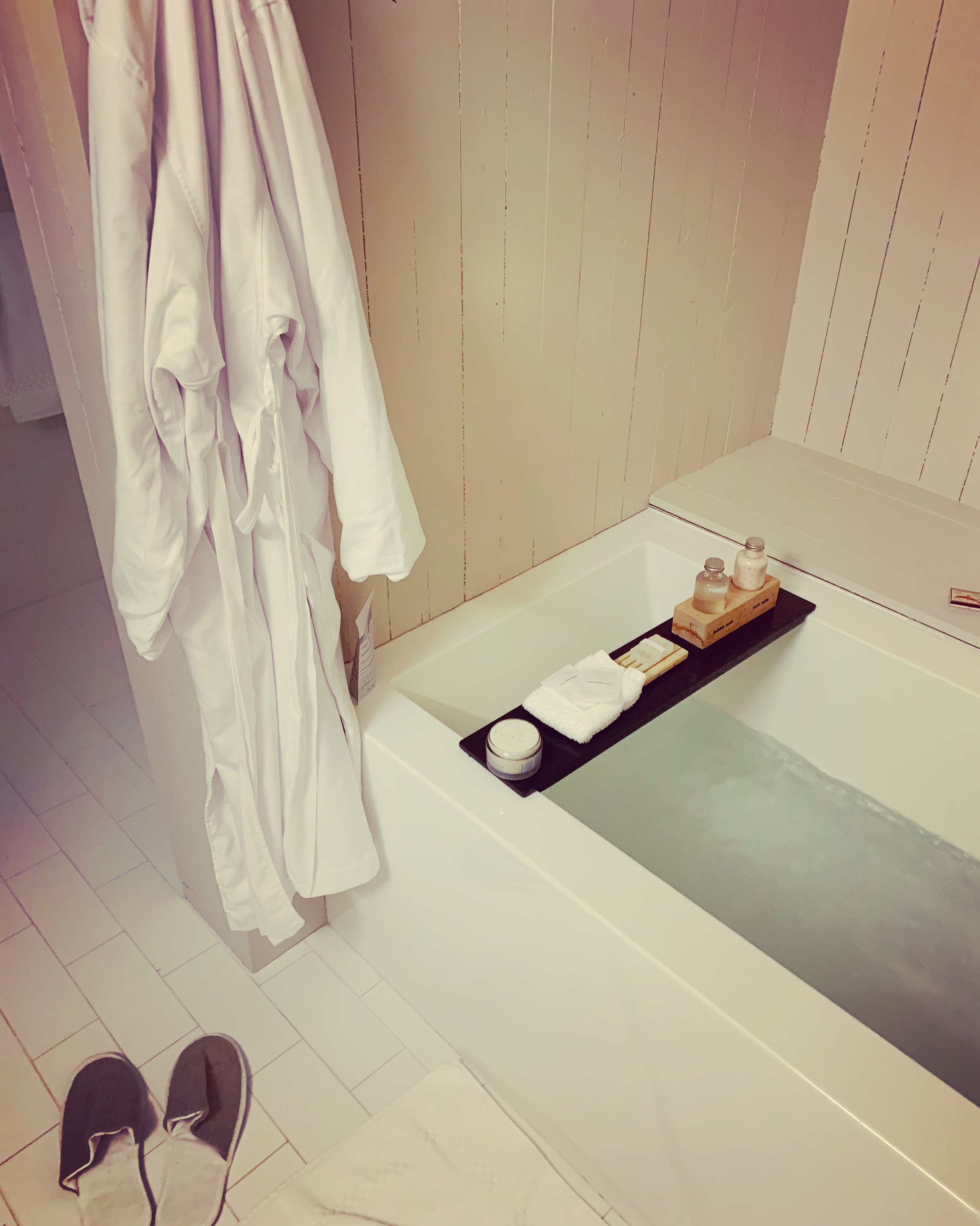 View of a bathroom with a bathtub, two bottles of toiletries, a towel, and bath products on a black tray. Part of the person's feet in sneakers is visible at the bottom of the image.