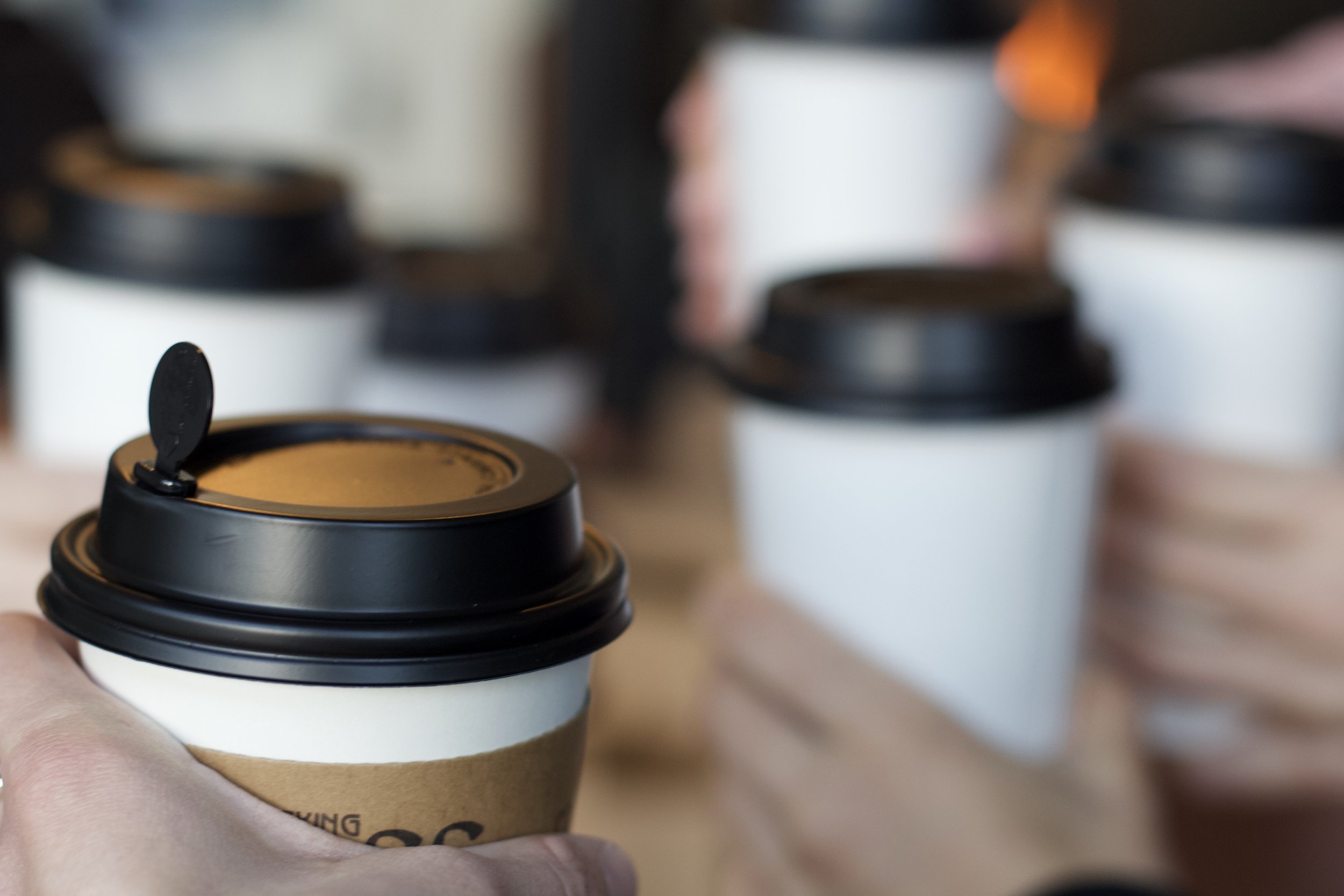 A person's hand holding a disposable coffee cup with a black plastic lid, with several similar cups in the background.