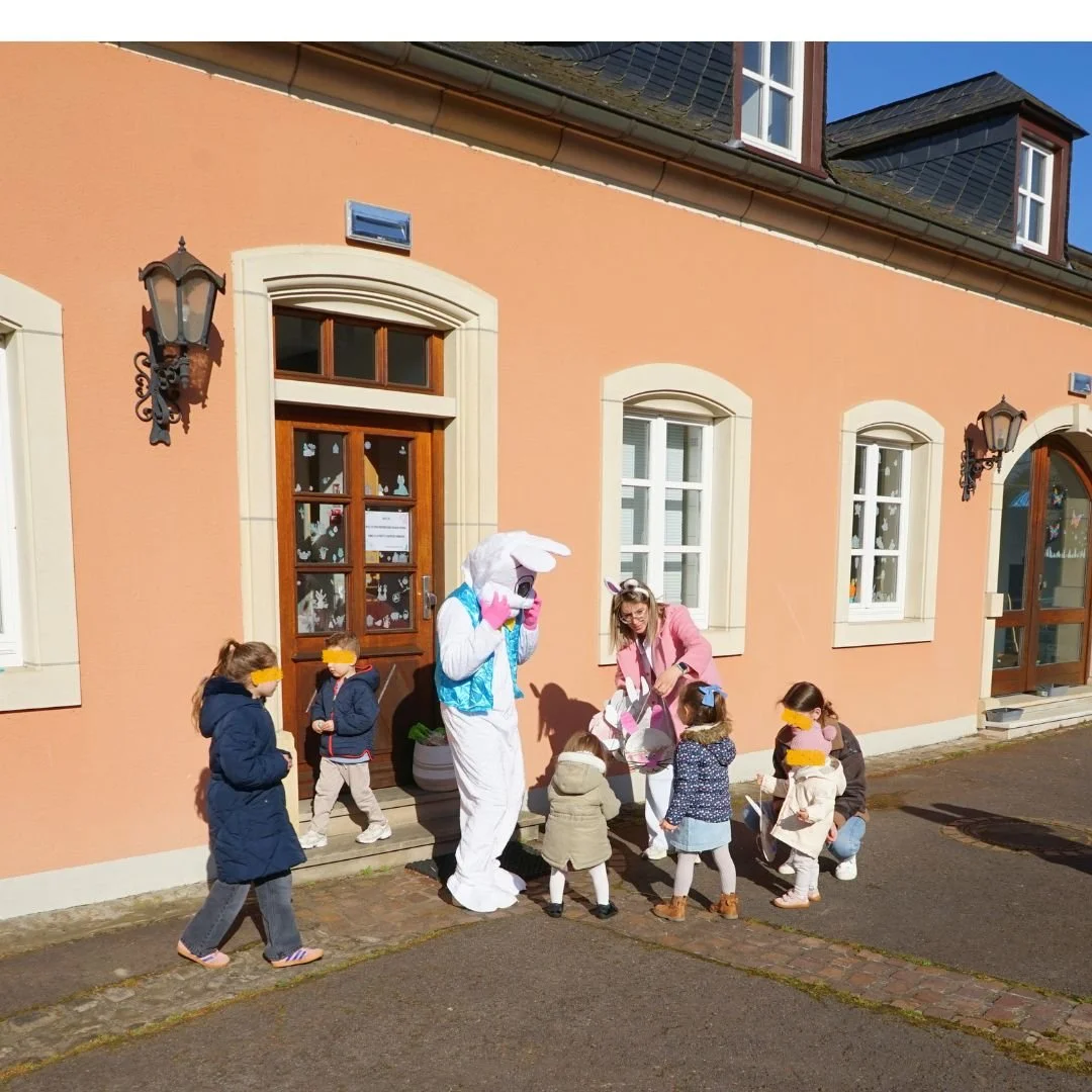 Le grand Lapin de Pâques en costume (gilet bleu) se tient près d'une éducatrice en veste rose devant l'entrée à porte en bois de la crèche. Ils sont entourés de jeunes enfants curieux.