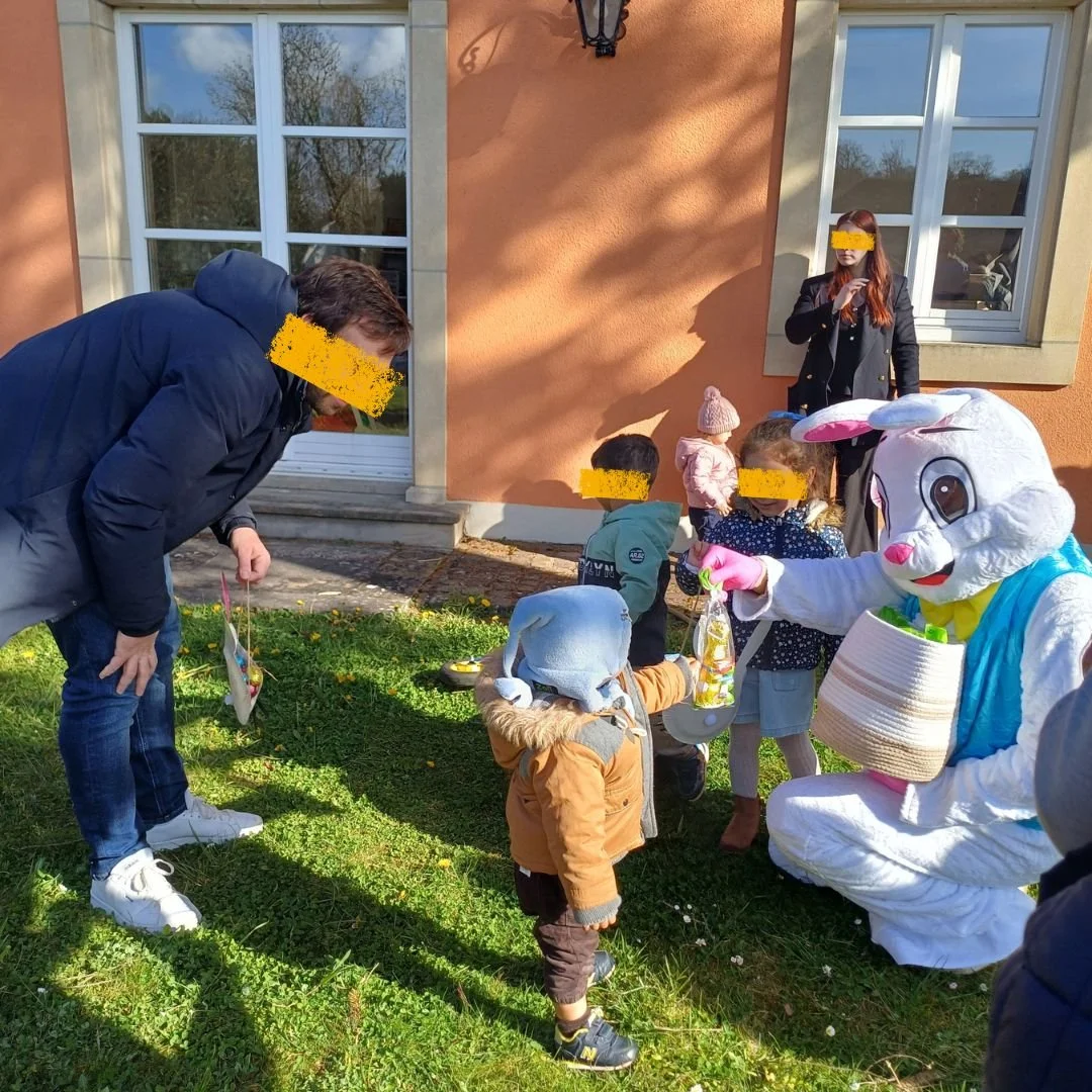 Le Lapin de Pâques à genoux distribue un sachet transparent rempli d'œufs en chocolat à un petit garçon portant un bonnet bleu, sous le regard bienveillant d'un père et d'autres enfants, dans le jardin ensoleillé de la crèche.