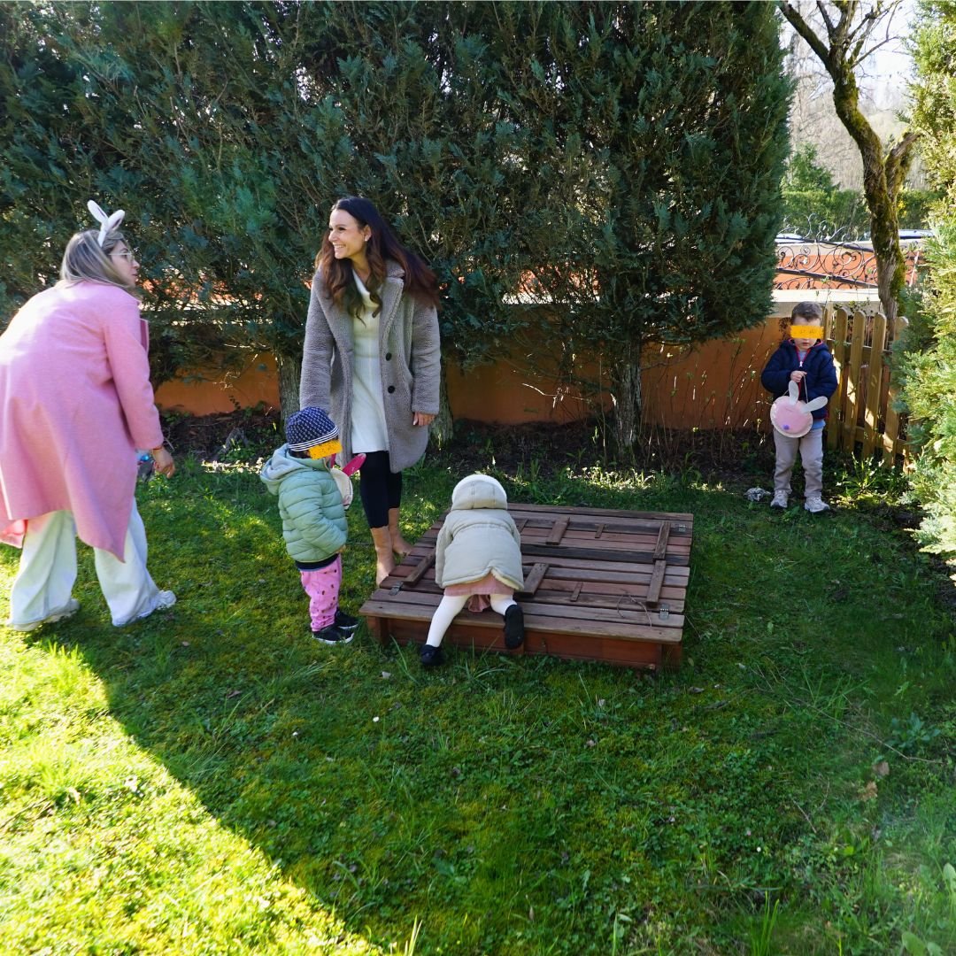 Deux jeunes enfants accompagnés de deux éducatrices (dont une portant un serre-tête oreilles de lapin) cherchent des œufs de Pâques dans un bac à sable en bois couvert dans un jardin vert.