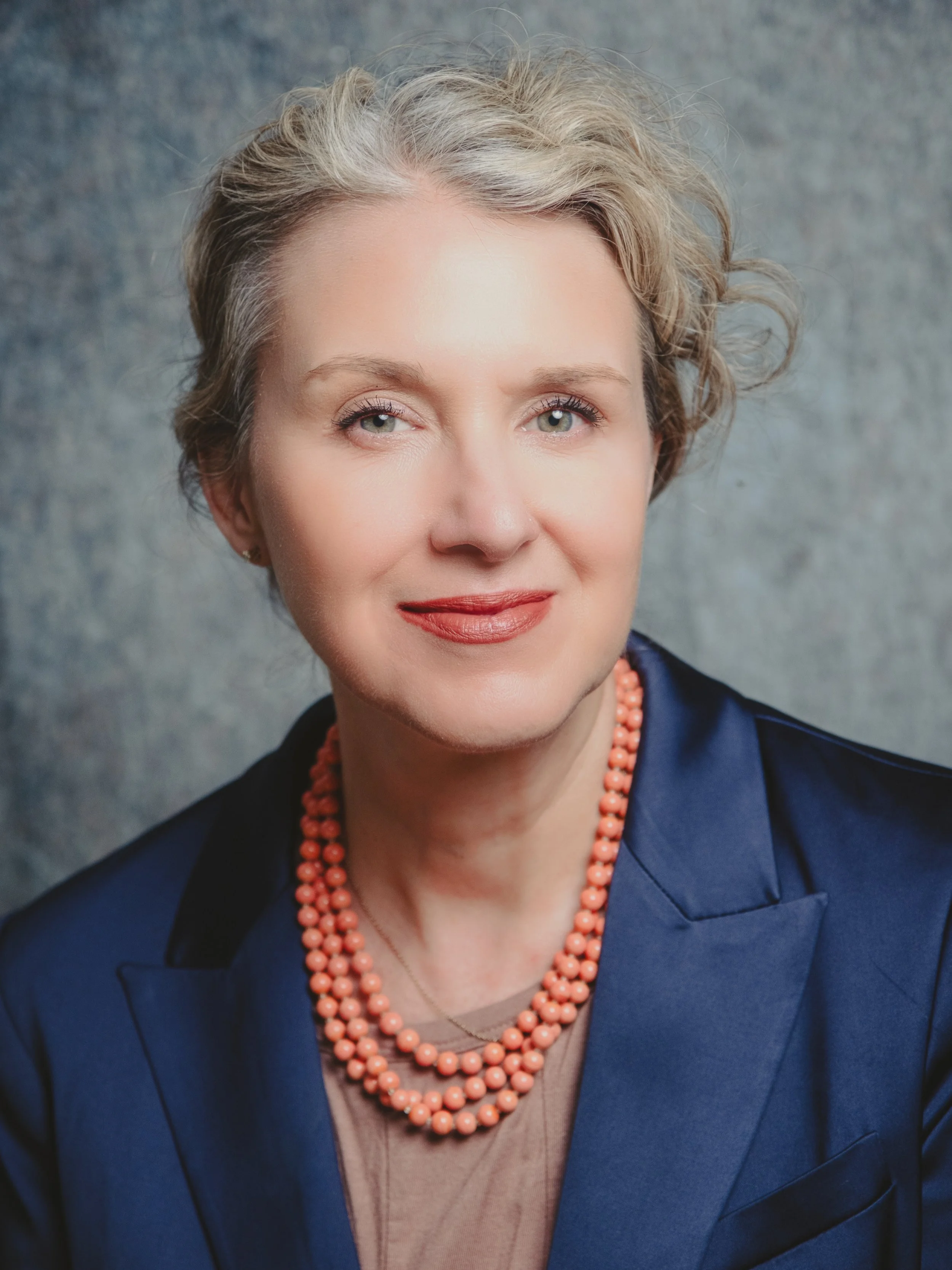 Portrait of a woman with short gray hair, wearing a blue blazer and coral beaded necklace.