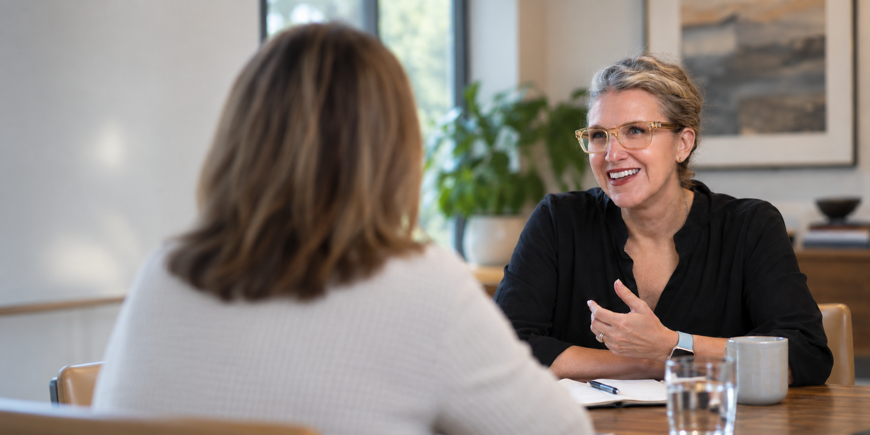 Two women having a conversation at a table in a bright room with large windows and framed artwork, one woman smiling and wearing glasses.