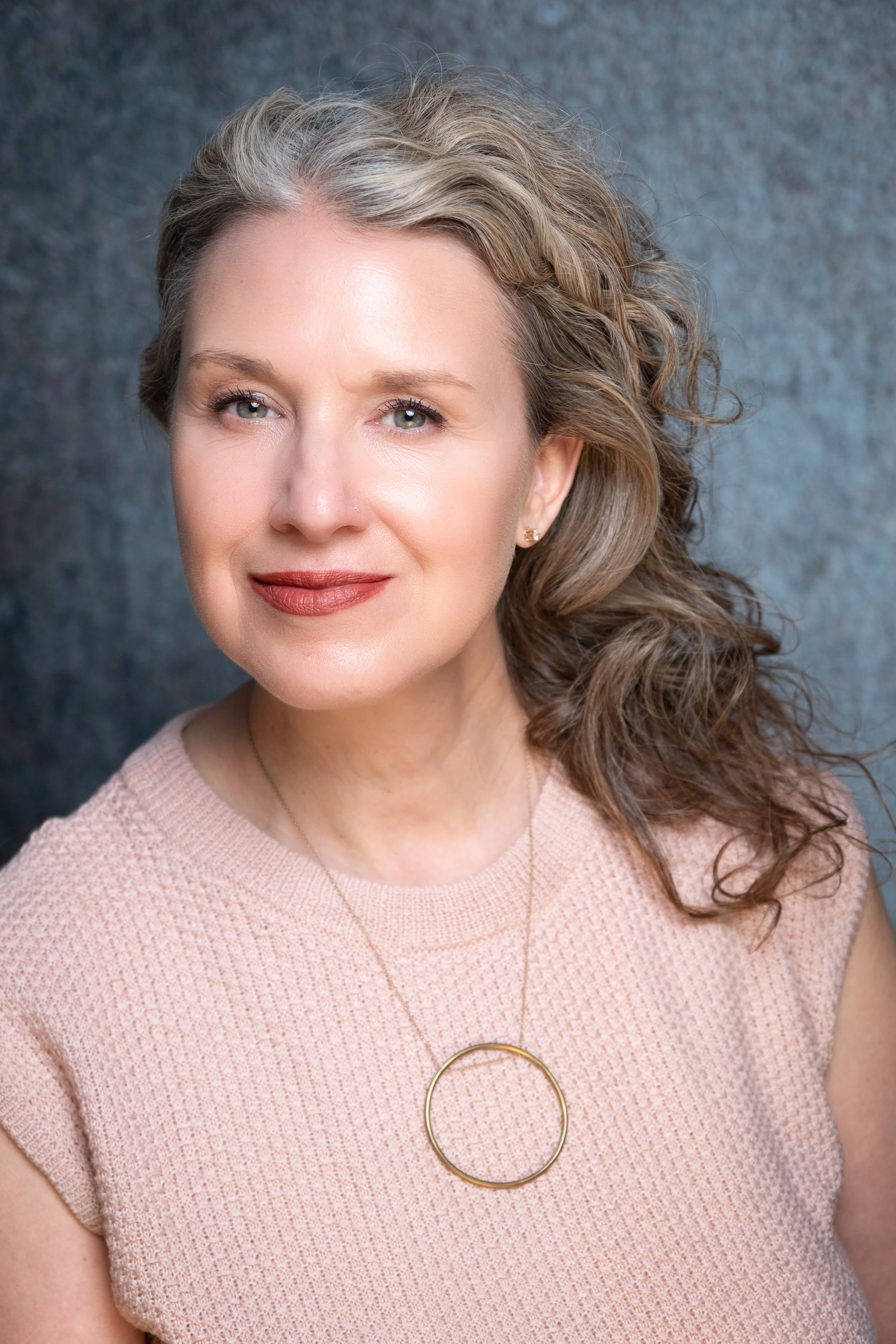 Woman with curly hair wearing a pink sweater and gold circle necklace, smiling against a textured gray background.