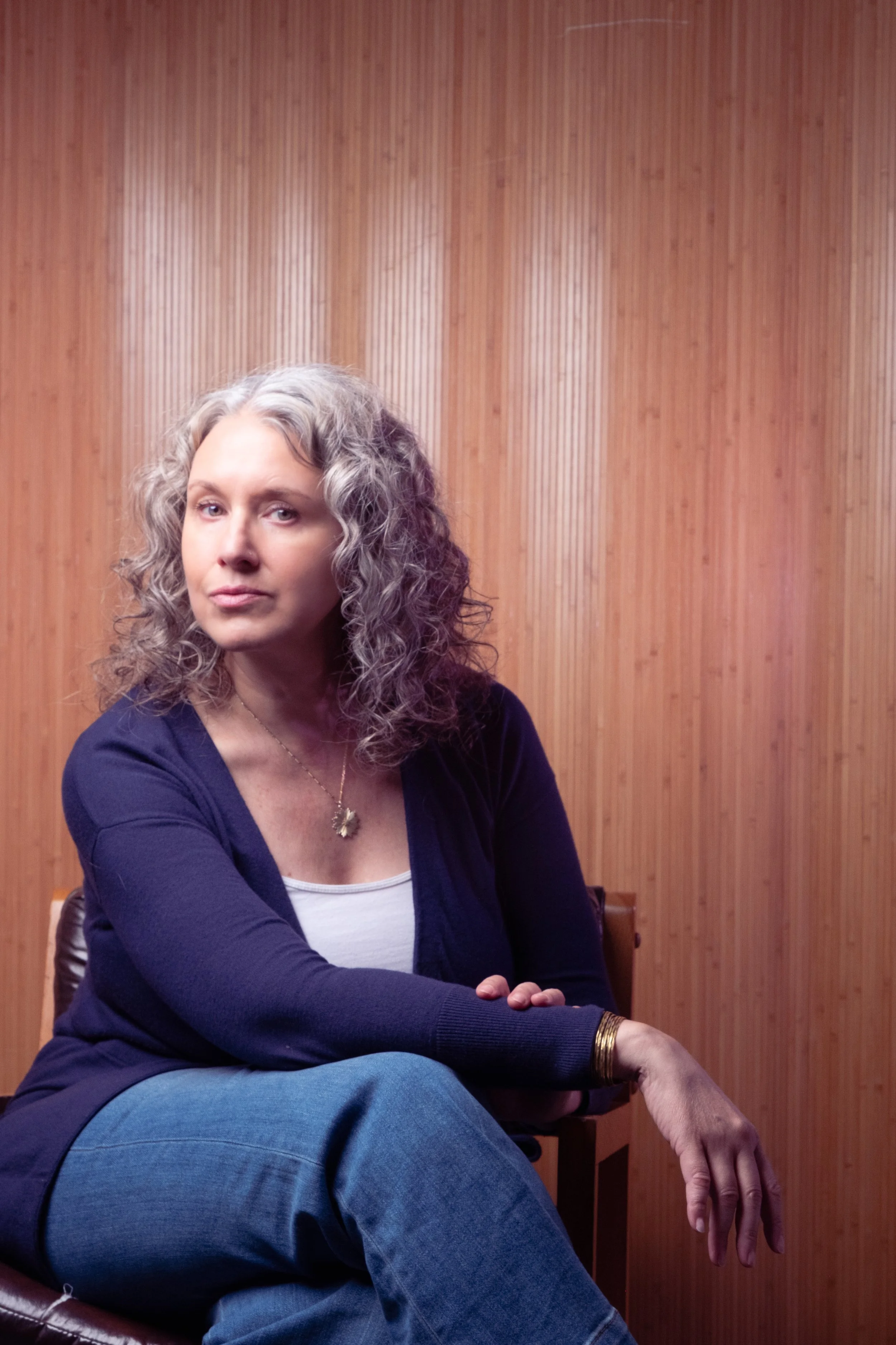 A woman with curly gray hair sitting in front of a wood-paneled wall, wearing a navy blue cardigan over a white top, with a gold necklace, a gold bracelet, and a gold ring, looking to the side.