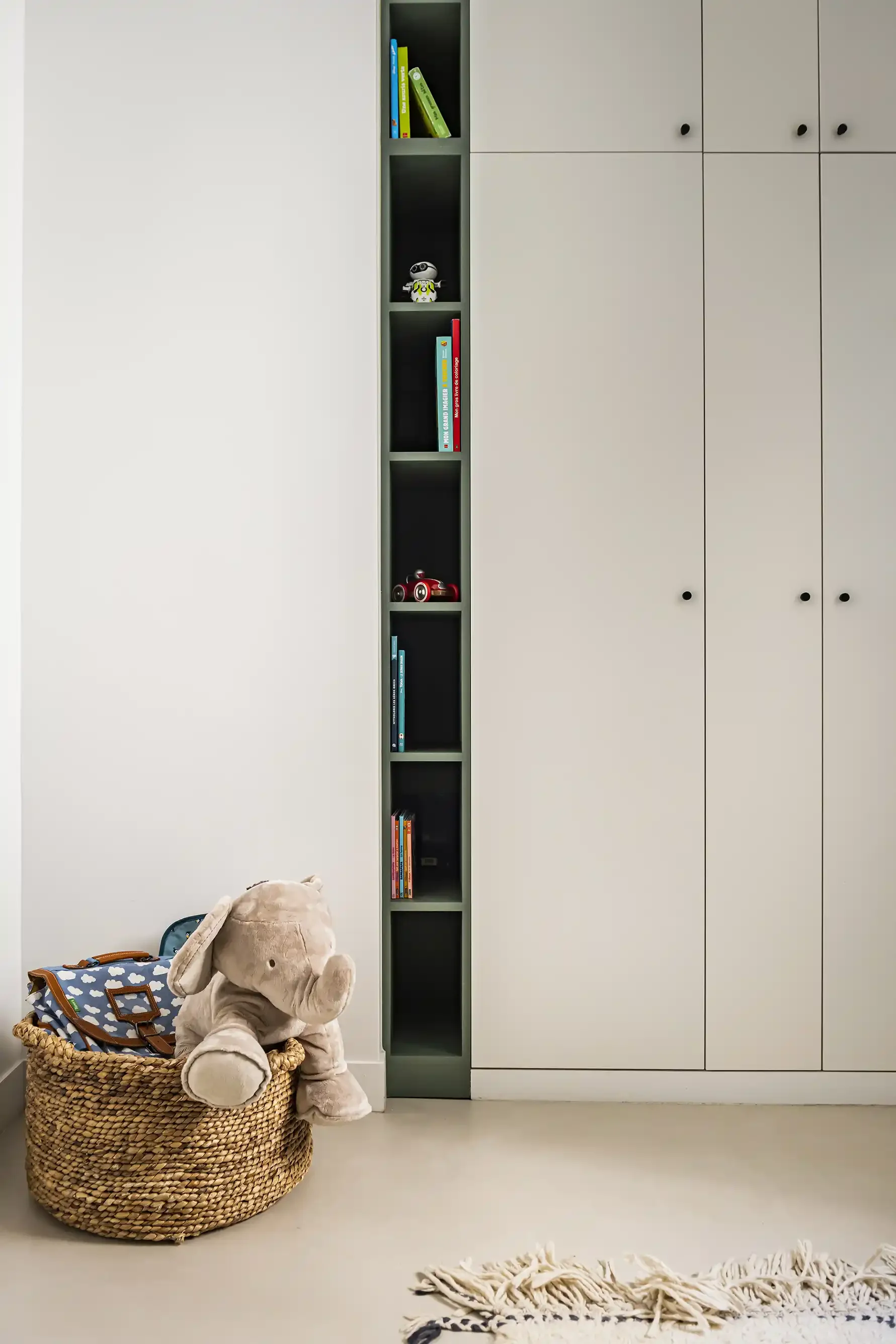 Zoom sur une bibliothèque encastrée vert sauge pour chambre d'enfant avec jouets, livres et un grand panier en osier contenant une peluche éléphant.