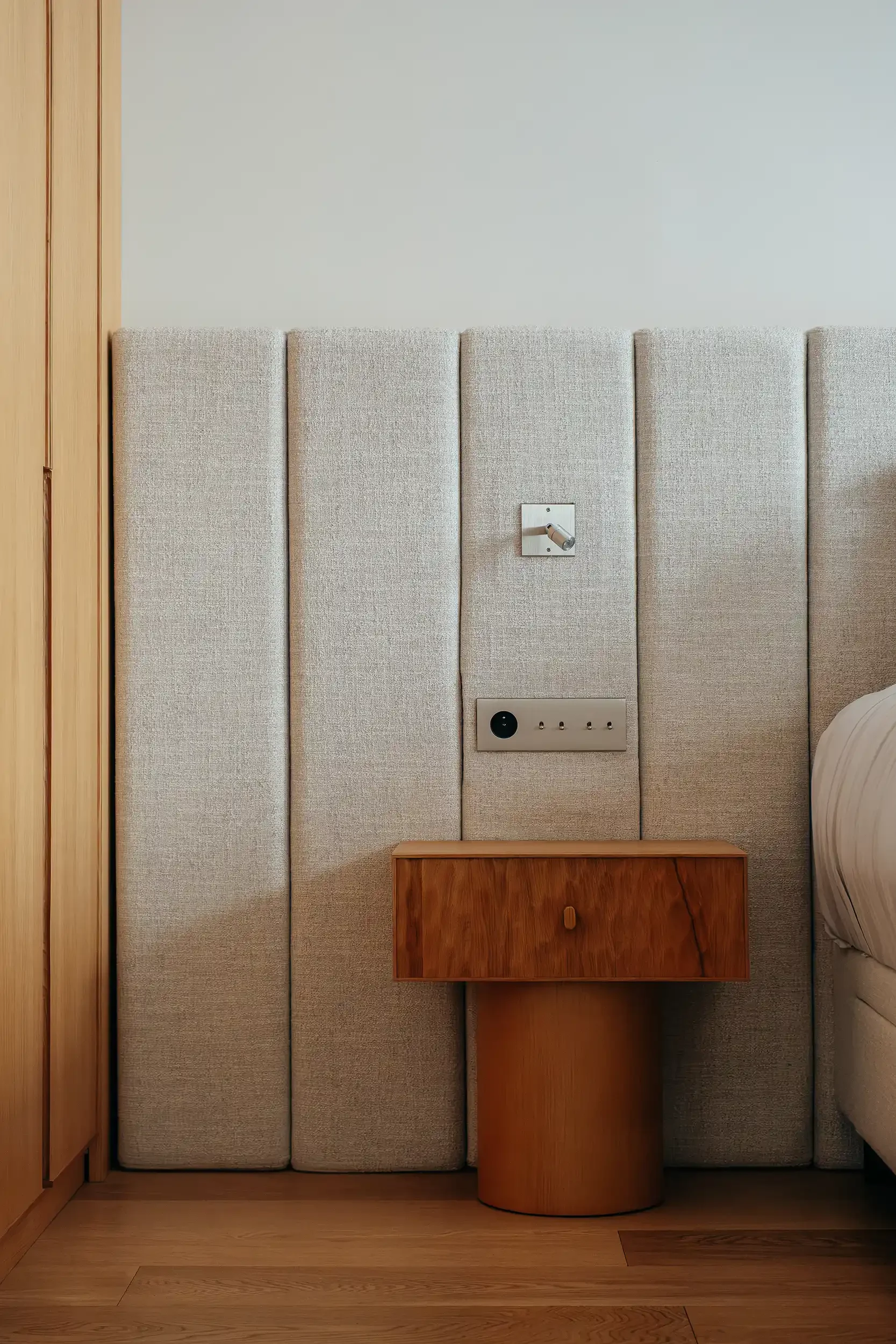 Détail de chambre avec table de chevet en bois minimaliste sur pied cylindrique et tête de lit capitonnée en tissu beige.