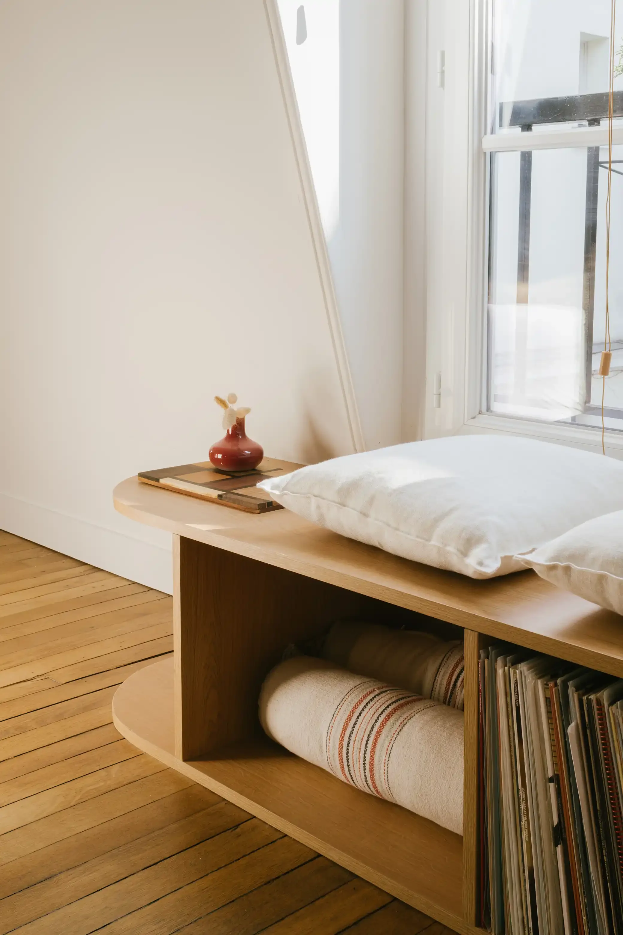 Banquette de lecture en bois sur-mesure installée sous une fenêtre avec niches de rangement pour disques vinyles et coussins blancs.