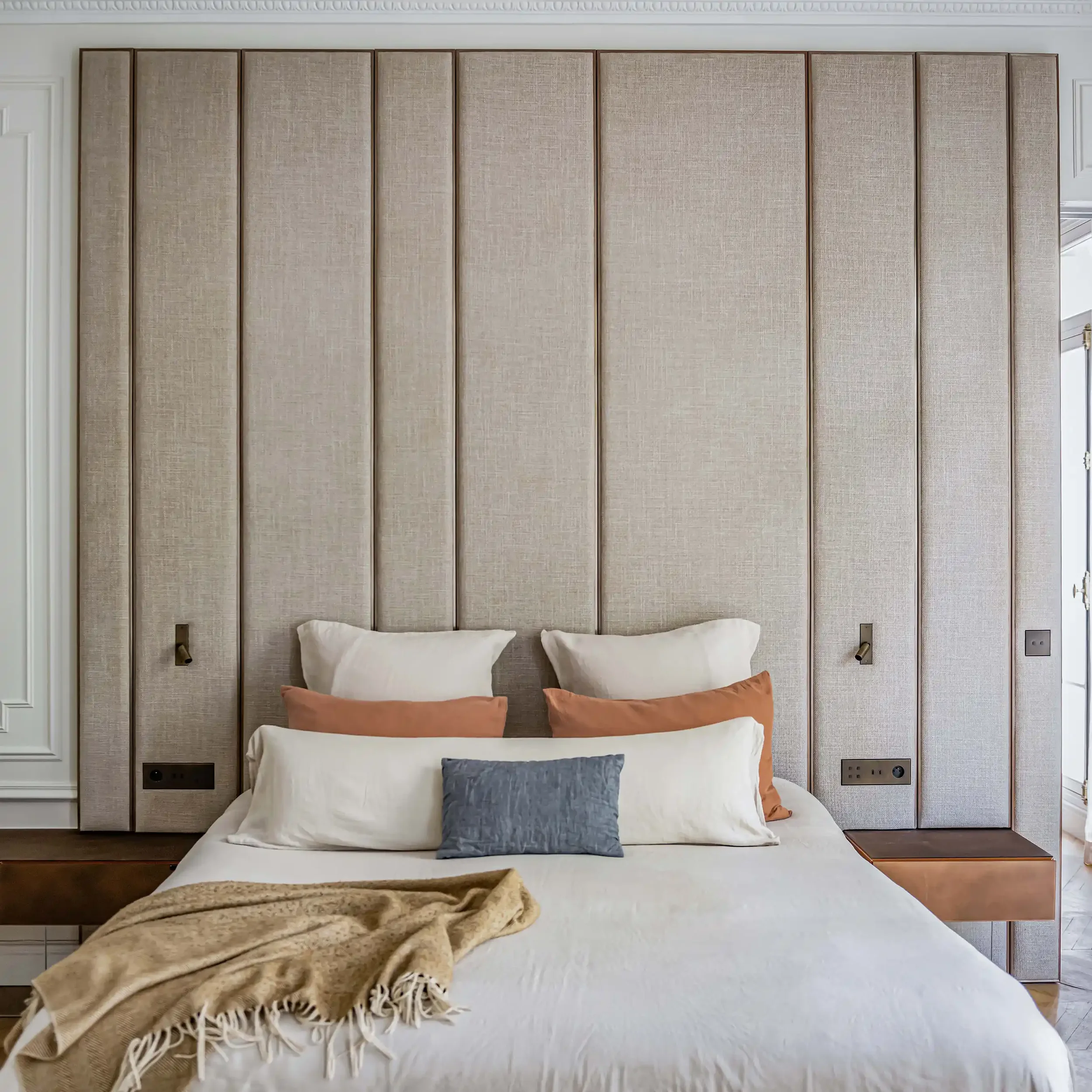 Tête de lit monumentale rembourrée en textile beige avec inserts en laiton et liseuses intégrées pour chambre de luxe.