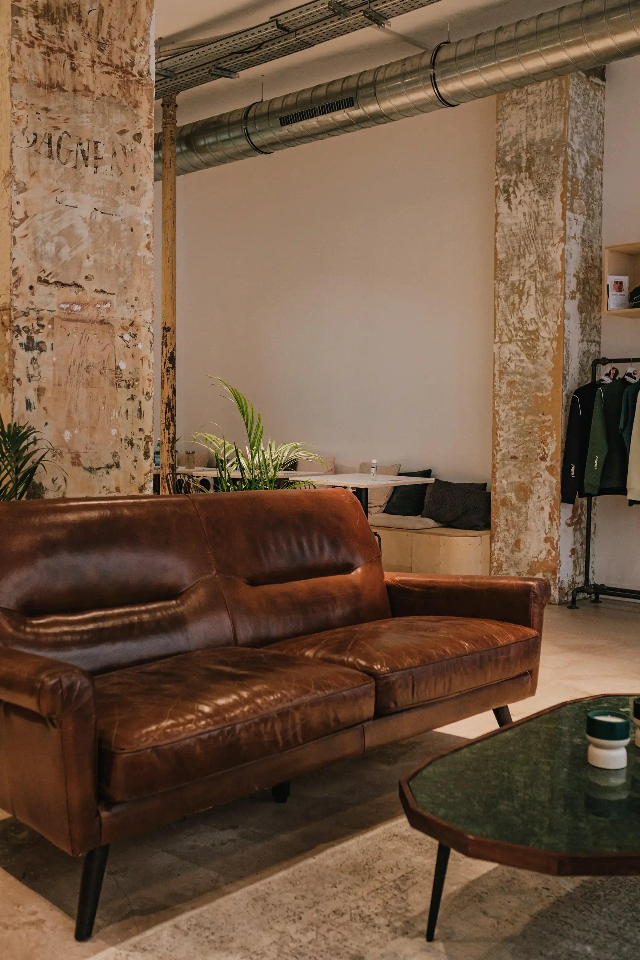 Salon de thé avec canapé en cuir vintage marron, table basse en pierre et colonnes en béton brut dans un espace au style industriel.