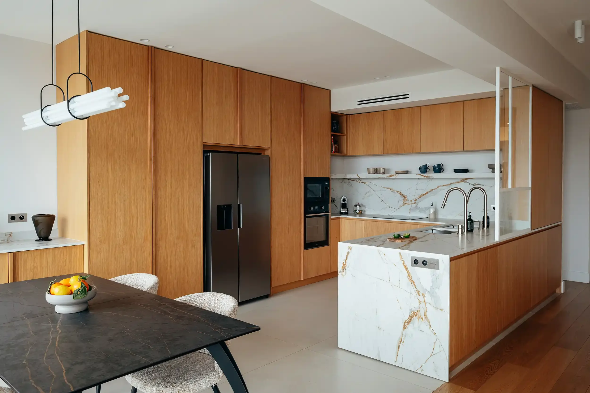 Vue d'ensemble d'une cuisine ouverte avec îlot central en marbre, table de salle à manger noire et luminaire design tubulaire.