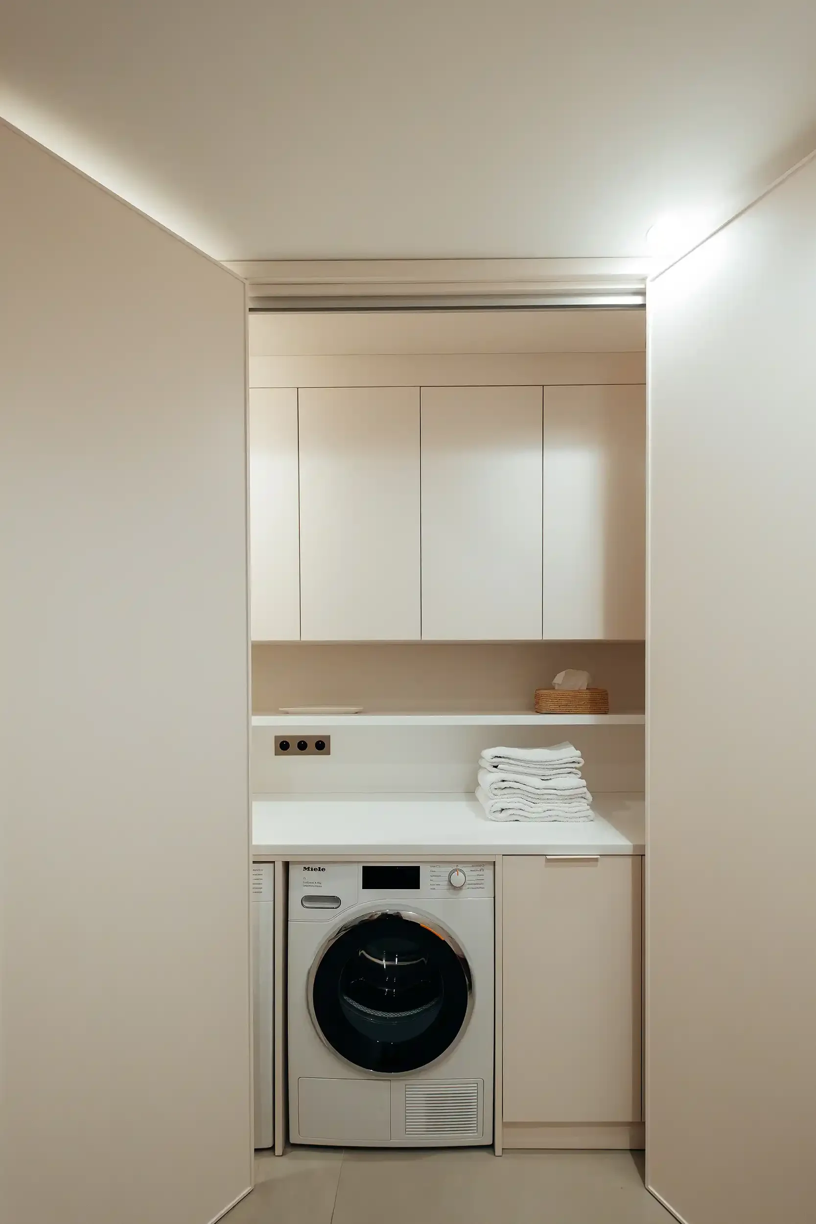Buanderie moderne escamotable avec lave-linge Miele intégré sous un plan de travail blanc et rangements toute hauteur.