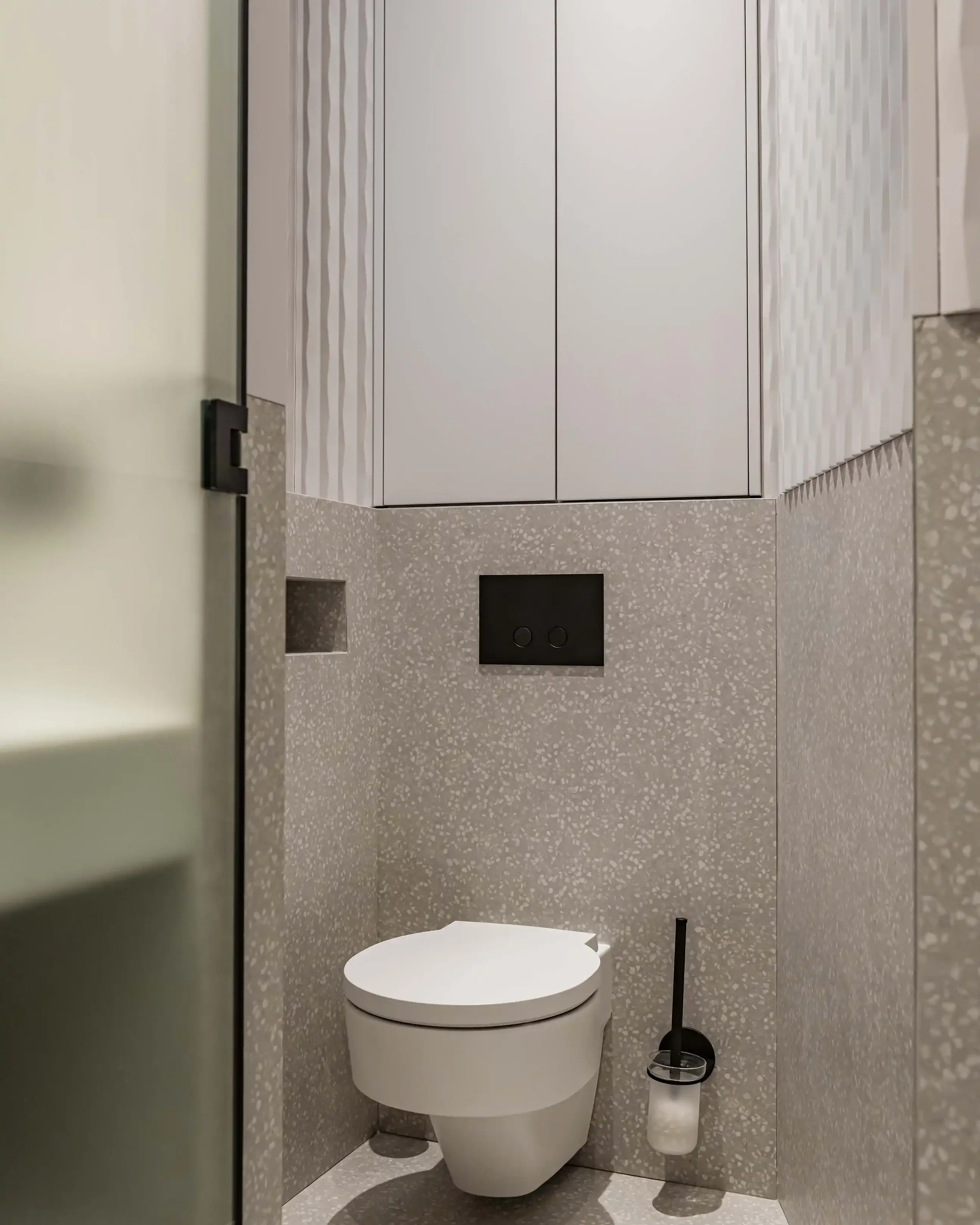 Toilettes suspendues blanches dans un cabinet moderne avec revêtement mural en terrazzo gris, niche intégrée et plaque de déclenchement noire mate.