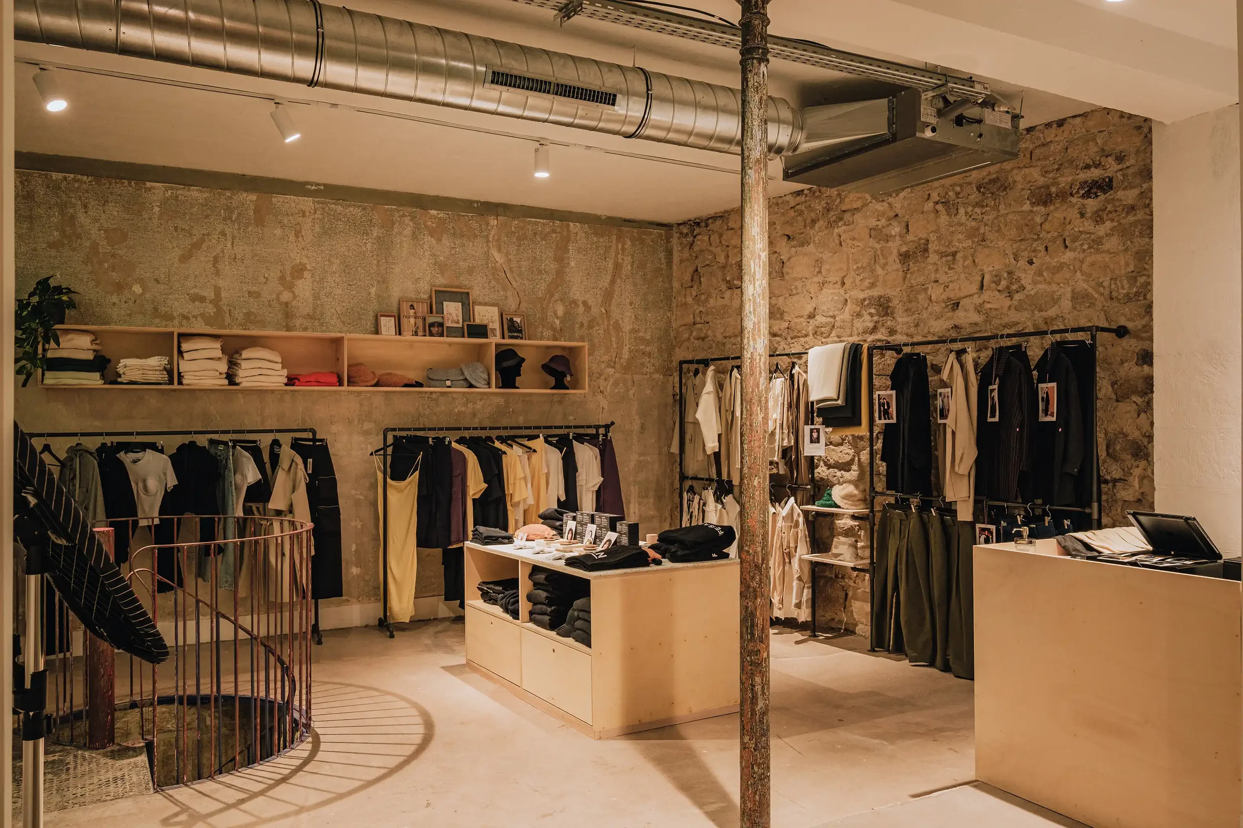 Boutique de mode avec vêtements suspendus sur rails industriels, mur en pierres apparentes et escalier en colimaçon design.